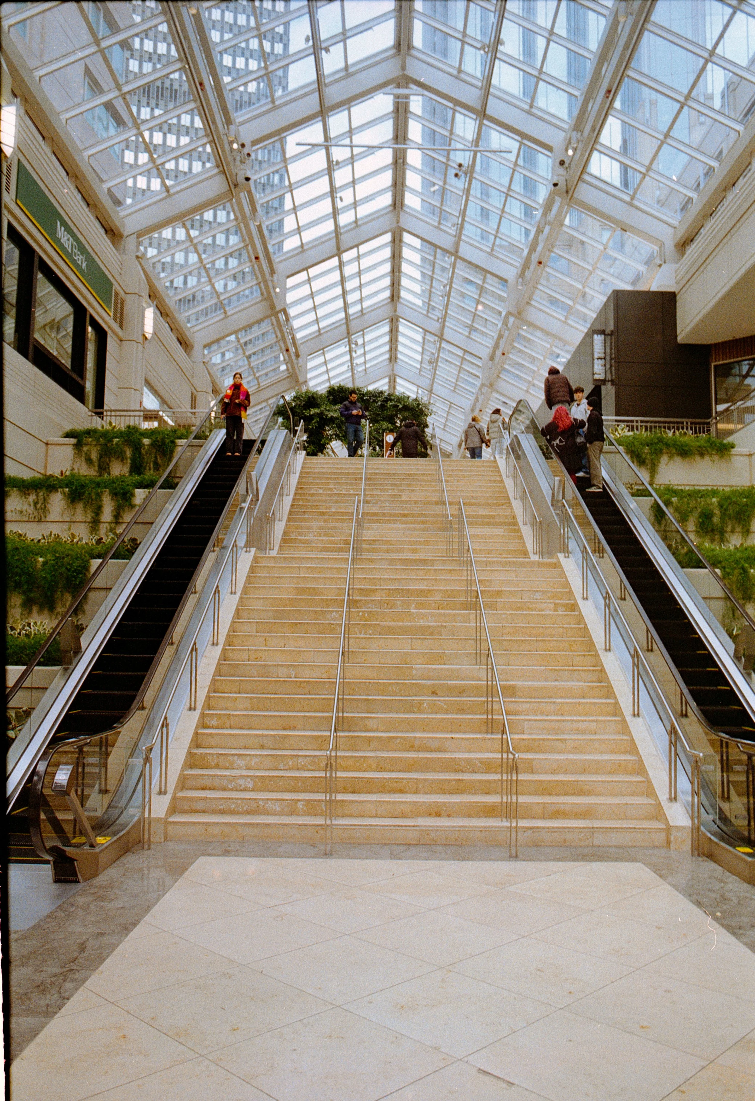 Indoor shopping mall with a large staircase in the center, flanked by escalators on both sides, glass ceiling, and people walking around.