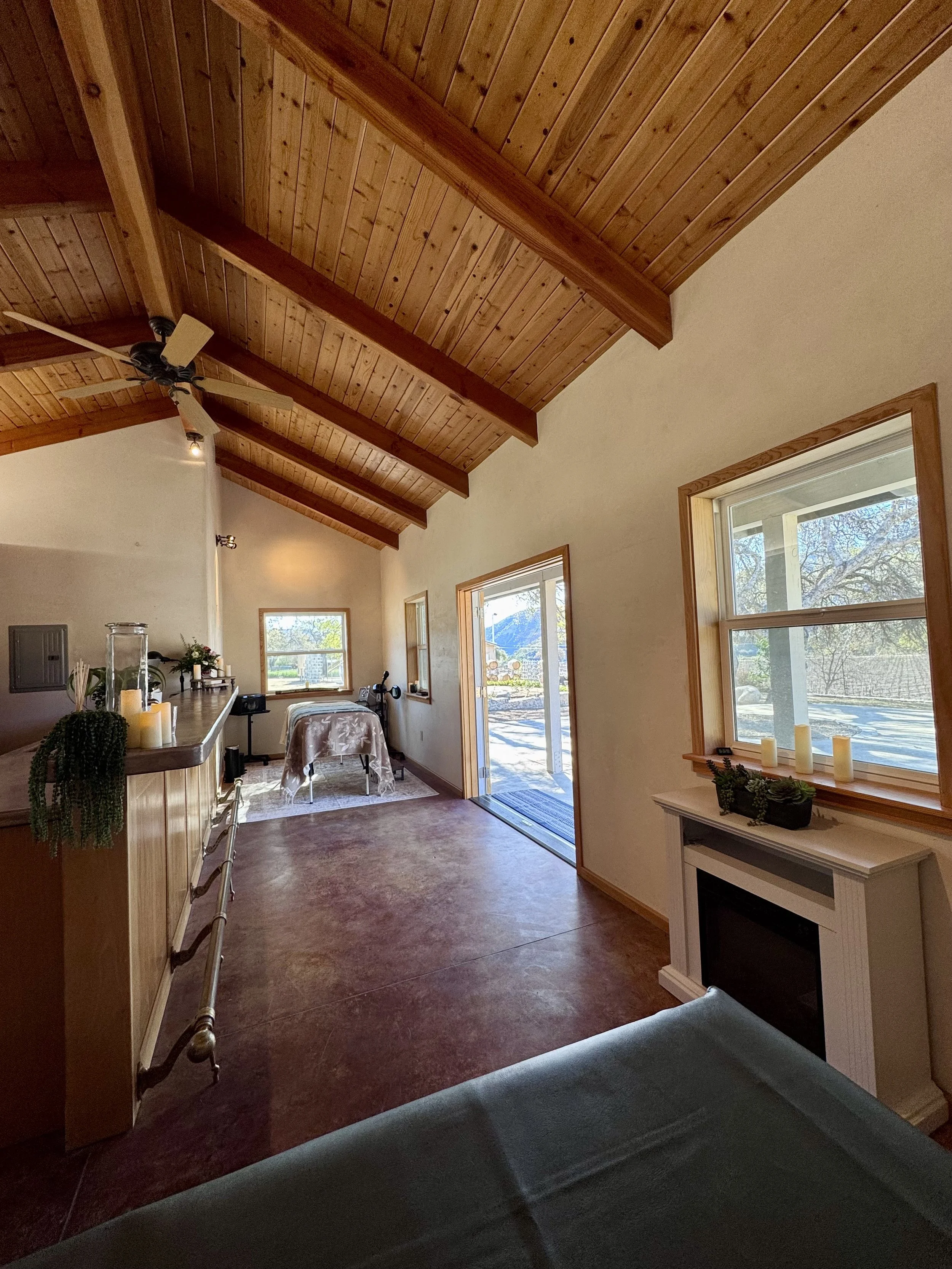 Interior of a room with a wood-paneled vaulted ceiling, a ceiling fan, and a doorway leading outside. The room has a window with candles and green plants on the white fireplace mantle. There is a table with a floral cover near the window, and a countertop with candles and plants along the wall.