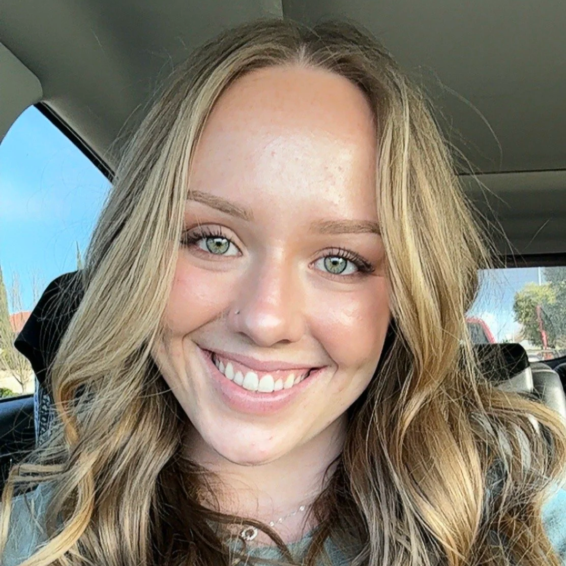 Close-up of a smiling woman with blonde hair and blue eyes inside a vehicle, with trees and blue sky visible outside the window.