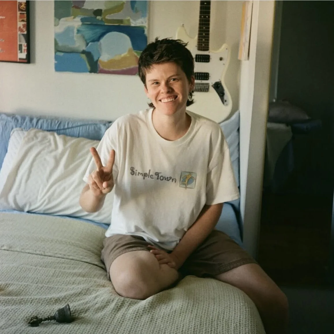 A person with short dark hair sitting on a bed, smiling, making a peace sign with their right hand, wearing a white "Simple Town" t-shirt and brown shorts, with a colorful abstract painting and a white guitar on the wall behind them.