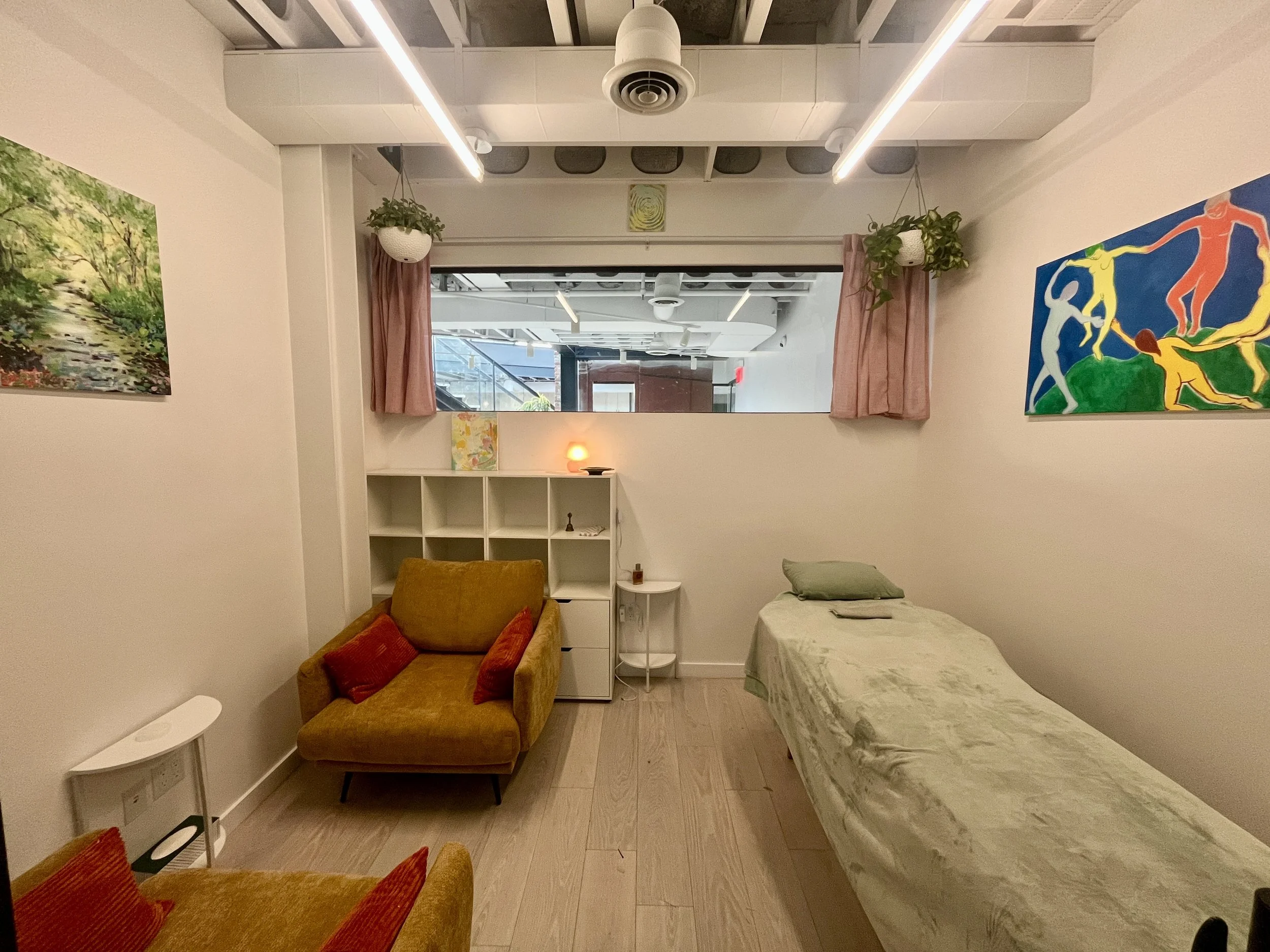 A small, cozy room with light-colored wooden flooring and white walls, featuring artwork on either side, a mustard yellow sofa with red pillows, a bed with a light green blanket, a window with pink curtains, and ceiling lights.