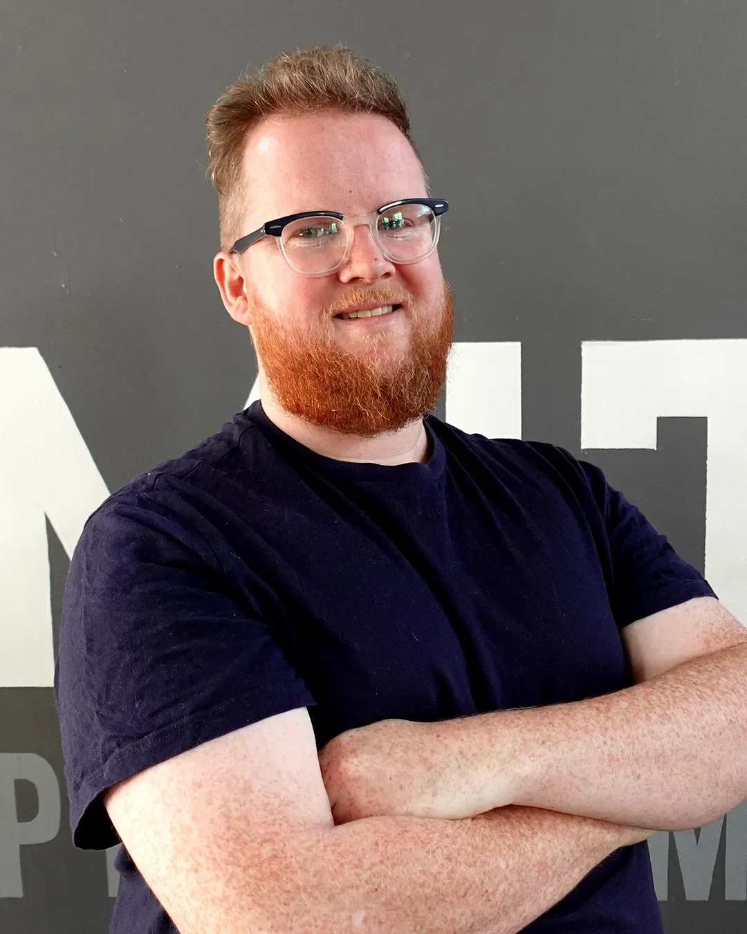 A man with glasses, a red beard, and short red hair smiling with arms crossed, standing in front of a dark gray wall with white text. Lewis Hutcheson Physio and Physiotherapist