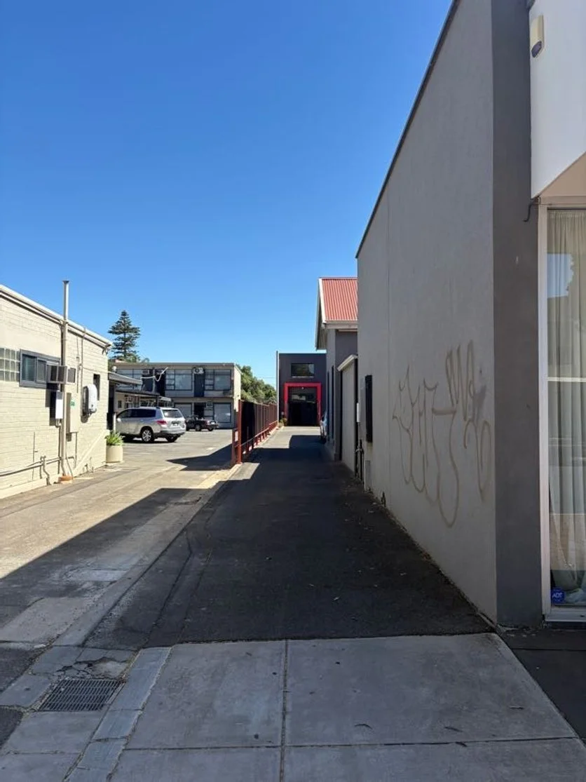 An alleyway between buildings with a clear blue sky. The sidewalk on the left side has parking spaces with cars, and the right side has a wall with graffiti. There is a small ramp and some steps at the end of the alley leading to a building with a red entrance.
