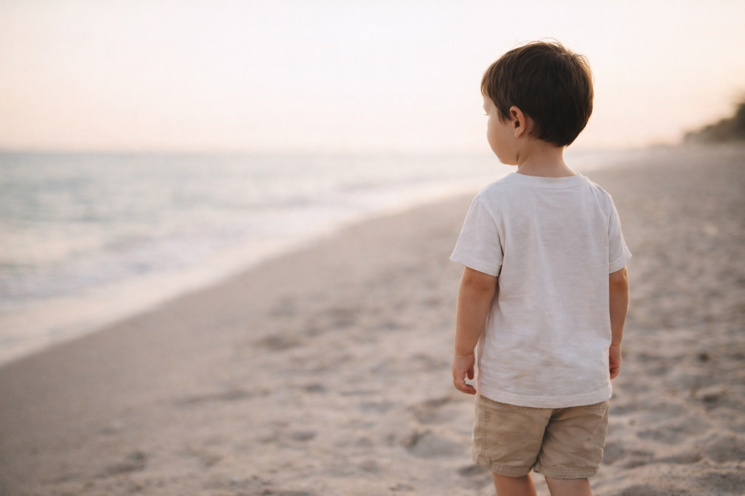 A young boy stands on a sandy beach, looking out at the ocean during sunset.