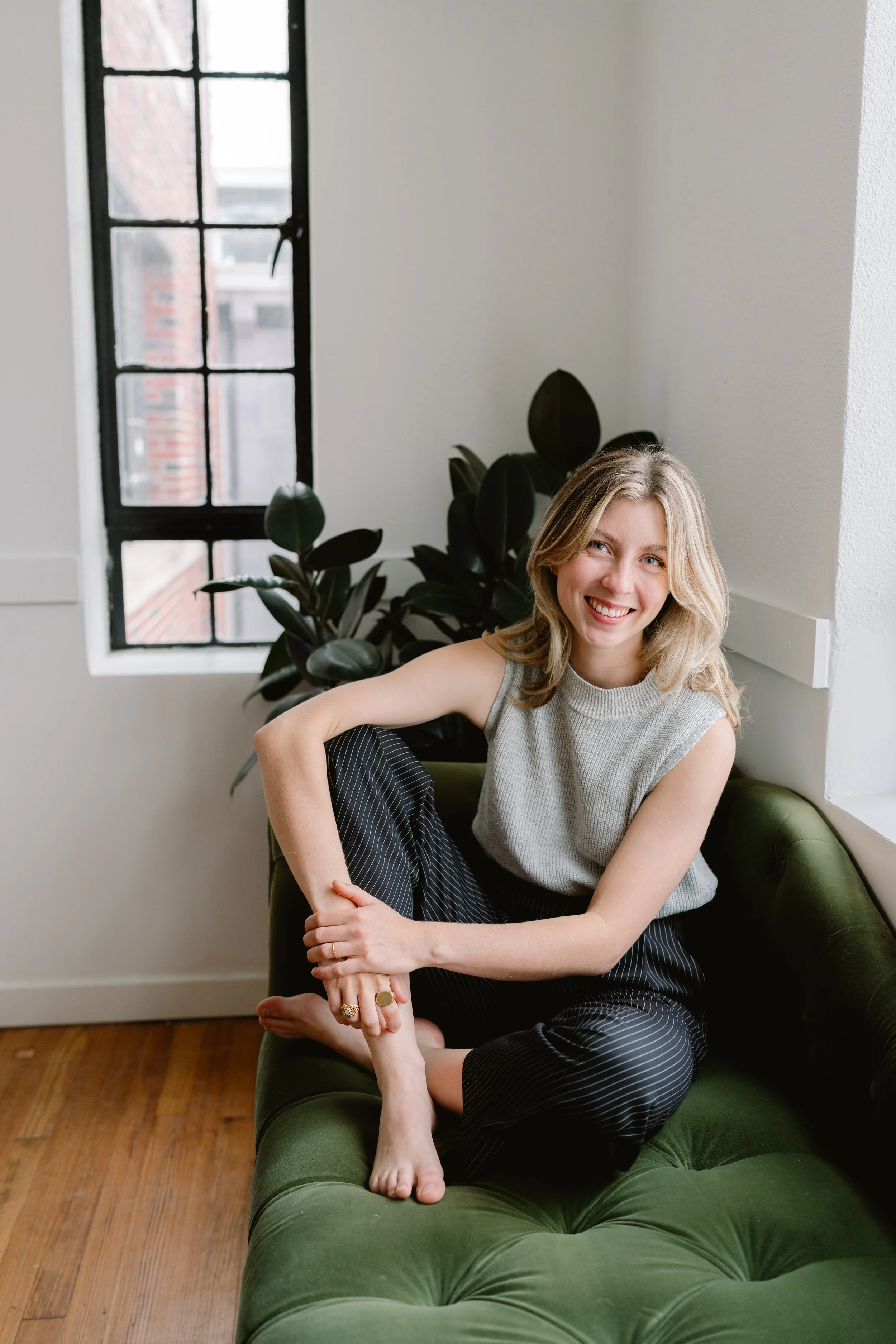 A young woman with blonde hair smiling and sitting cross-legged on a green sofa indoors with a black-framed window and indoor plants behind her.