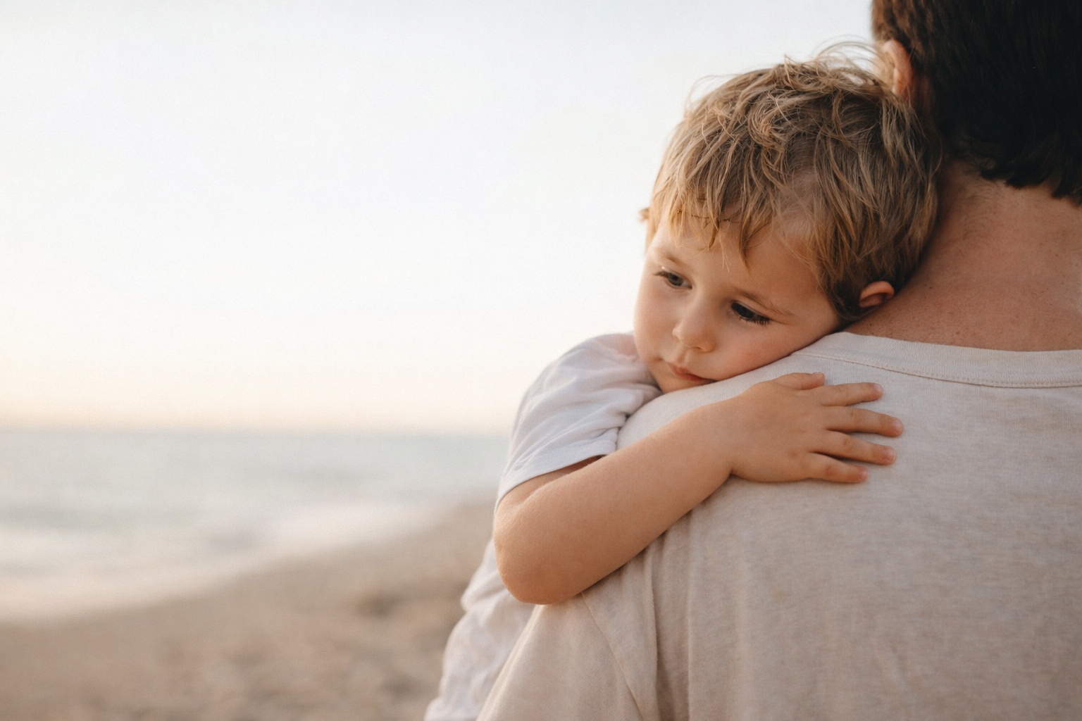 A young child with blonde hair resting on an adult's shoulder at the beach during sunset or early evening.
