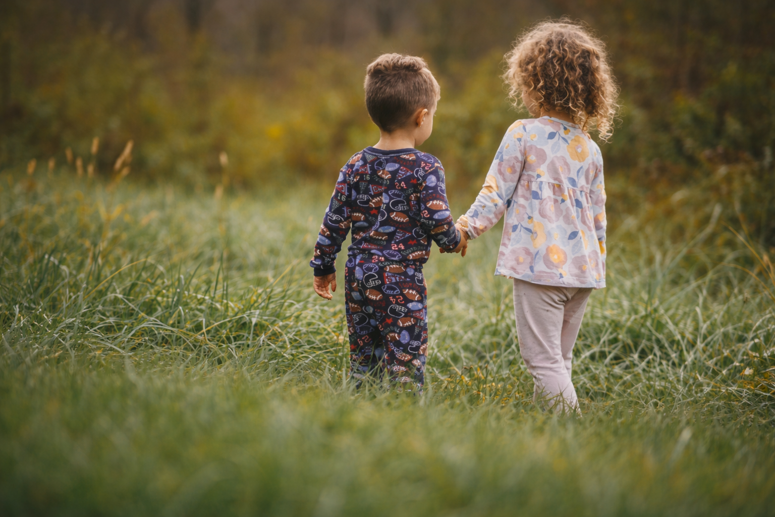 Two young children, a boy and a girl, holding hands and walking in a grassy field with trees in the background.