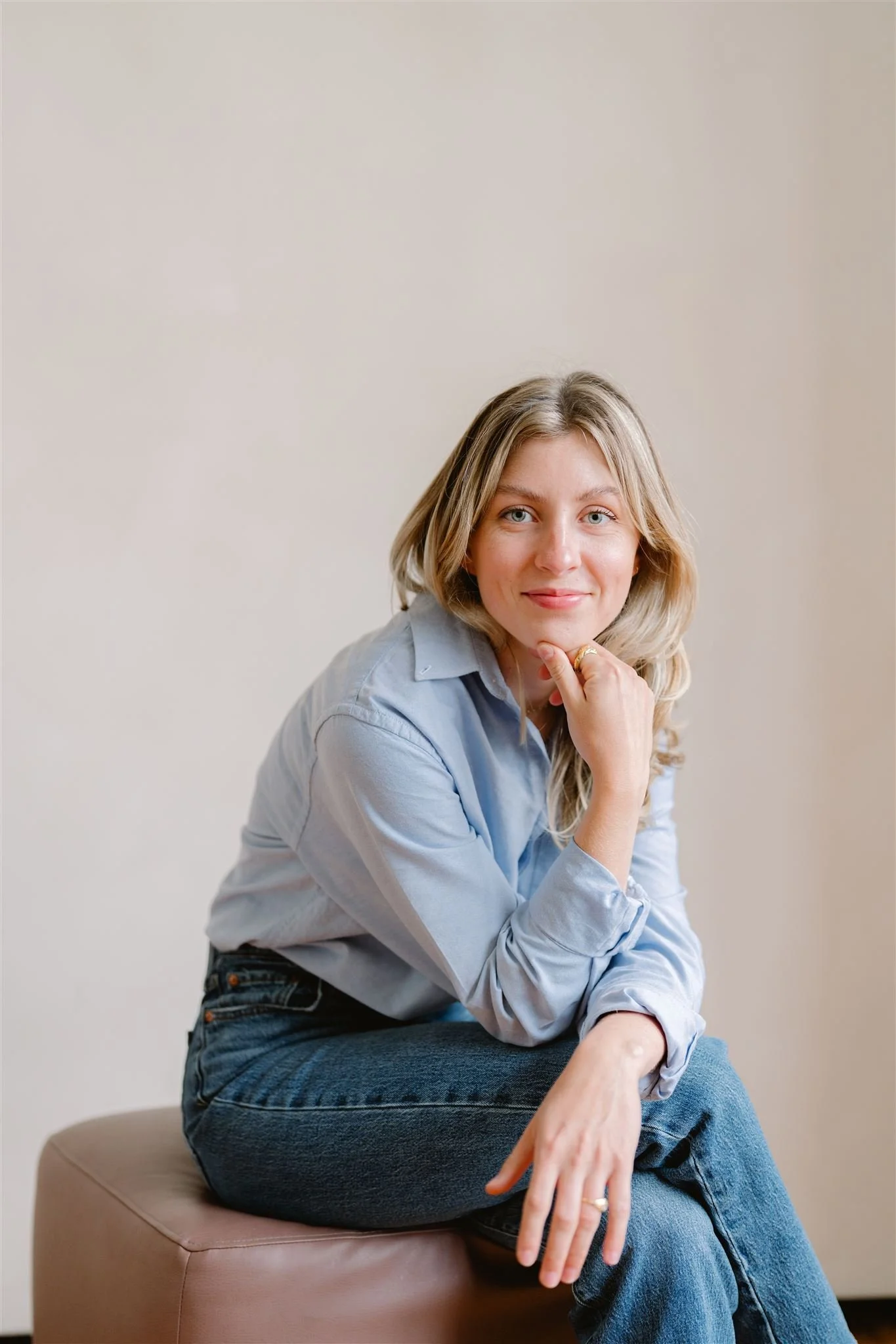 Portrait of a woman with blonde hair sitting on a beige ottoman against a plain wall, wearing a light blue shirt and blue jeans, smiling gently with her chin resting on her hand.