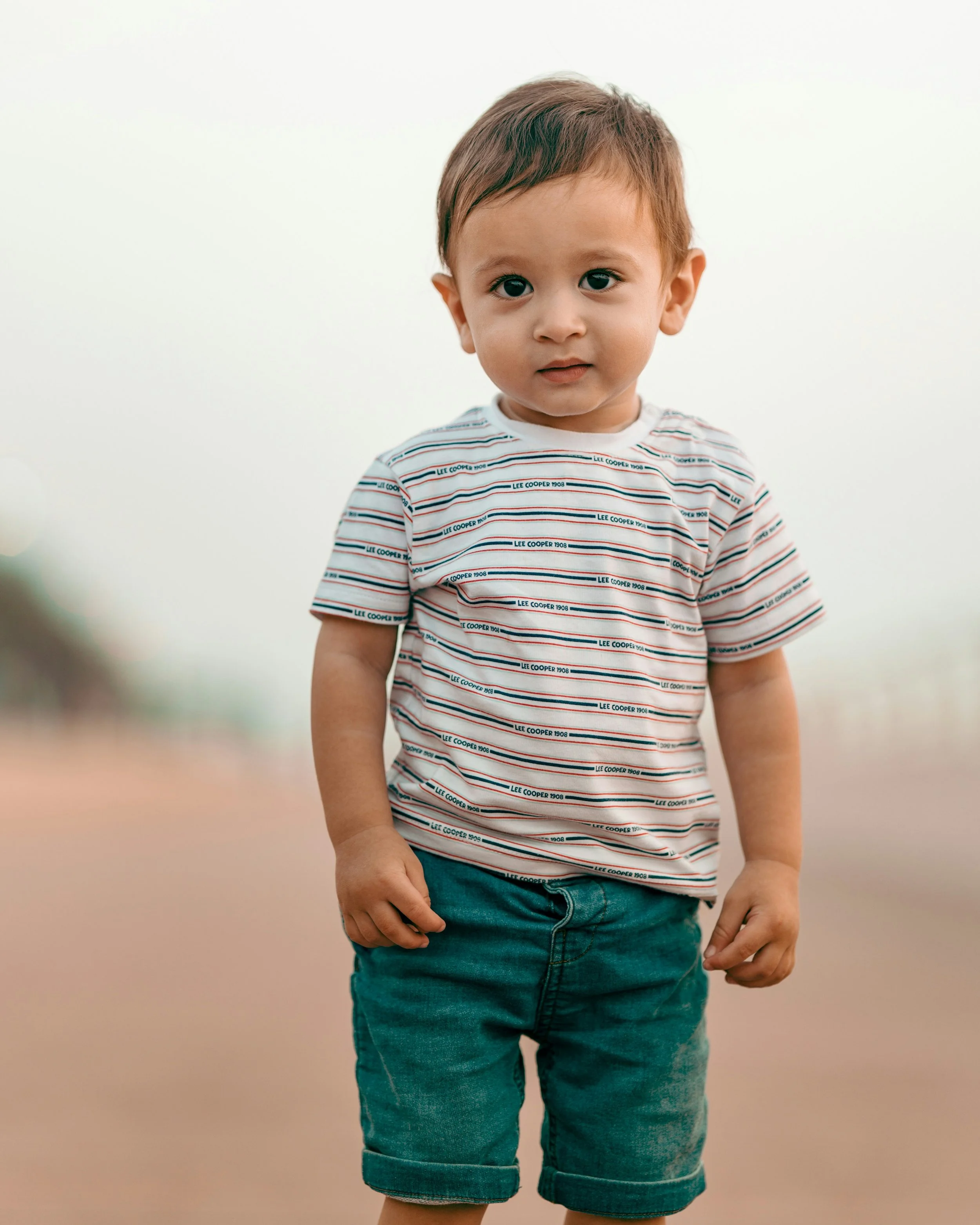 A young boy with brown hair and big eyes, wearing a white striped t-shirt with blue and red lines and denim shorts, standing outdoors on a hazy day.
