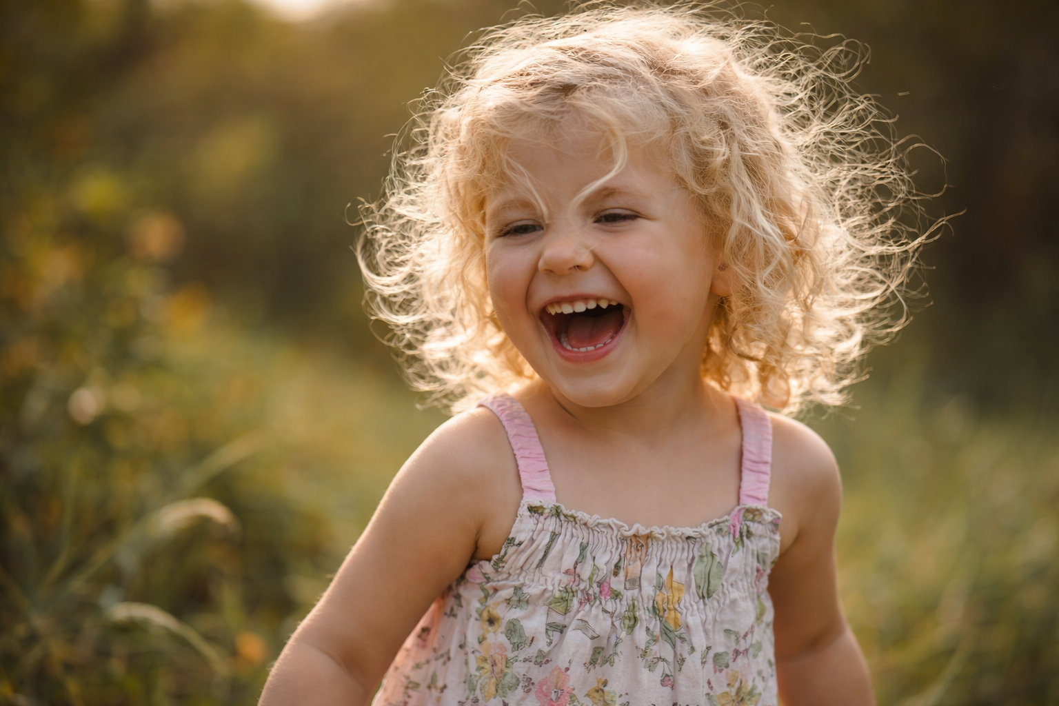 A young girl with blonde, curly hair laughing outdoors in natural light.