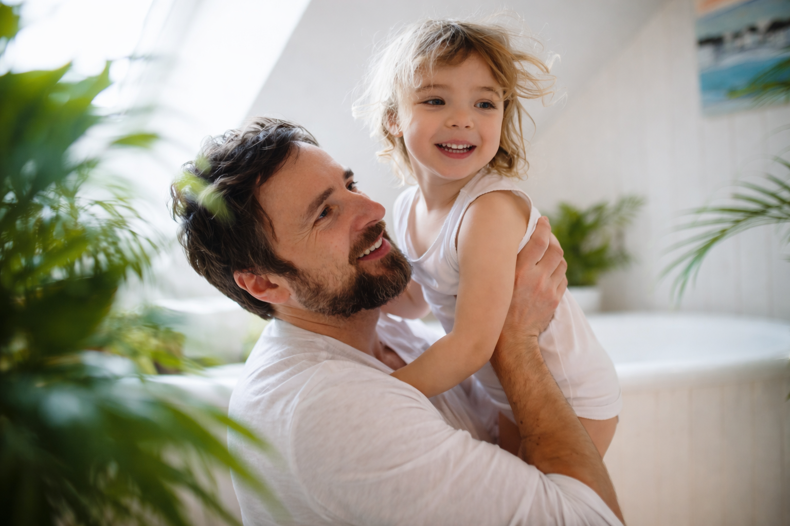 A man holding a smiling young girl, both looking happy, in a bright bathroom with plants in the background.