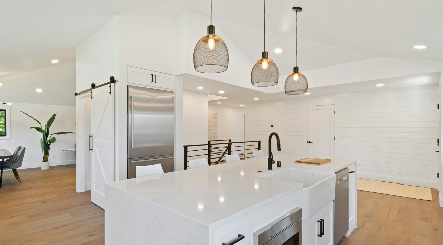 Modern open-concept kitchen with white cabinets, a large white island with a sink, black faucet, and three pendant lights, stainless steel refrigerator, hardwood floors, and a dining area with a potted plant in the background.