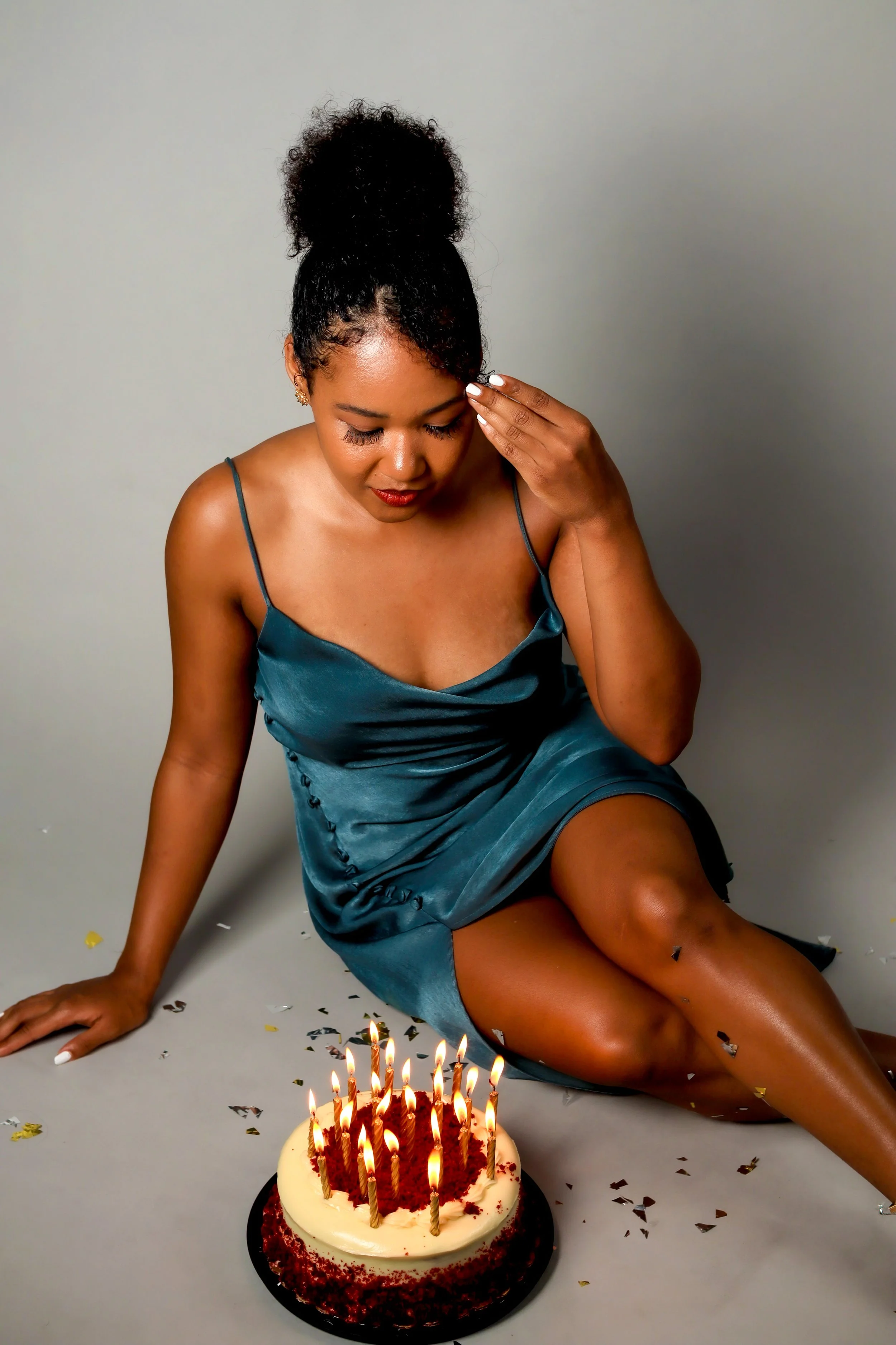 A woman in a teal dress sitting on the floor during a celebration with a birthday cake with lit candles and confetti on the floor.