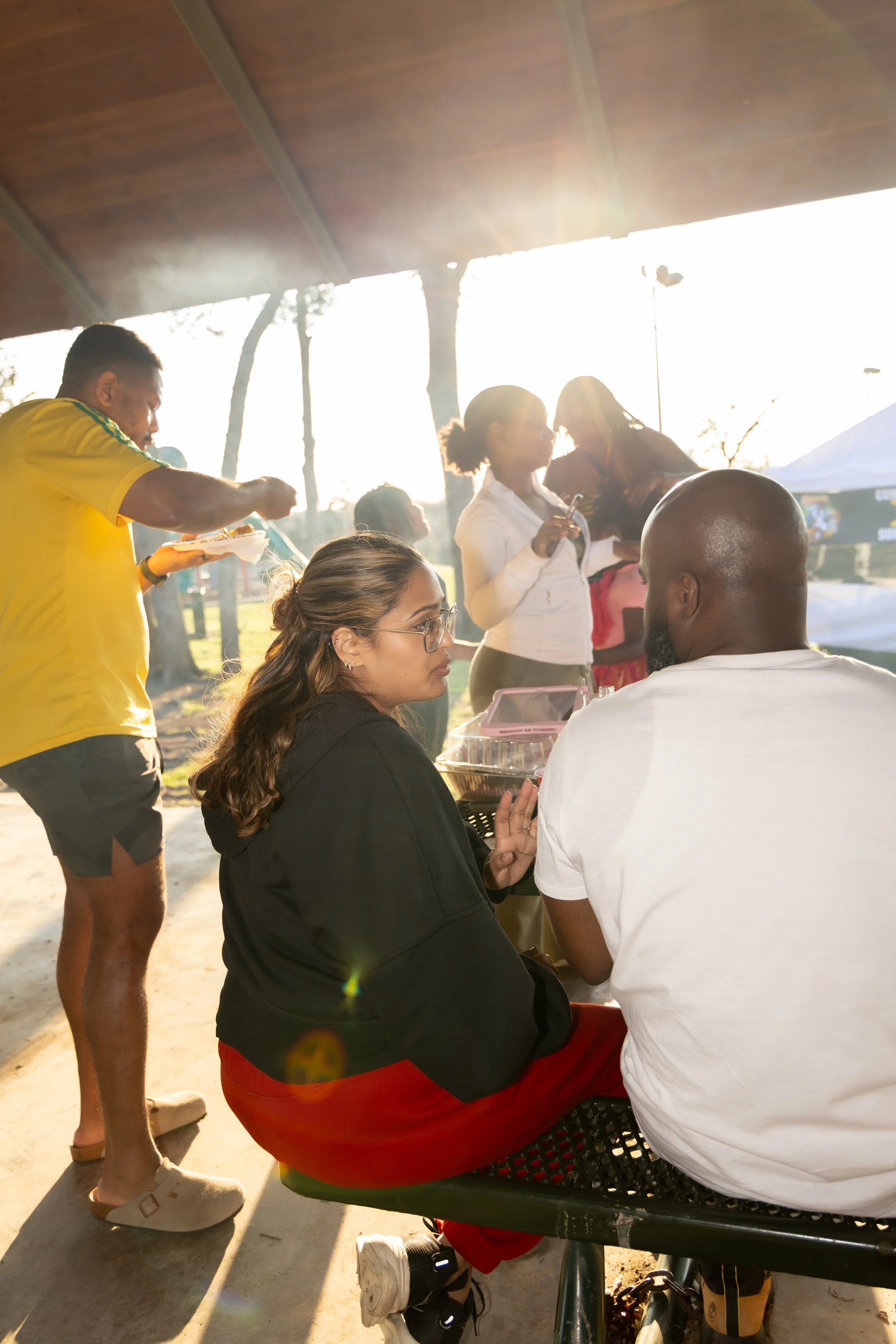 People gathering at an outdoor event during sunset, some seated and some standing, with food and drinks.