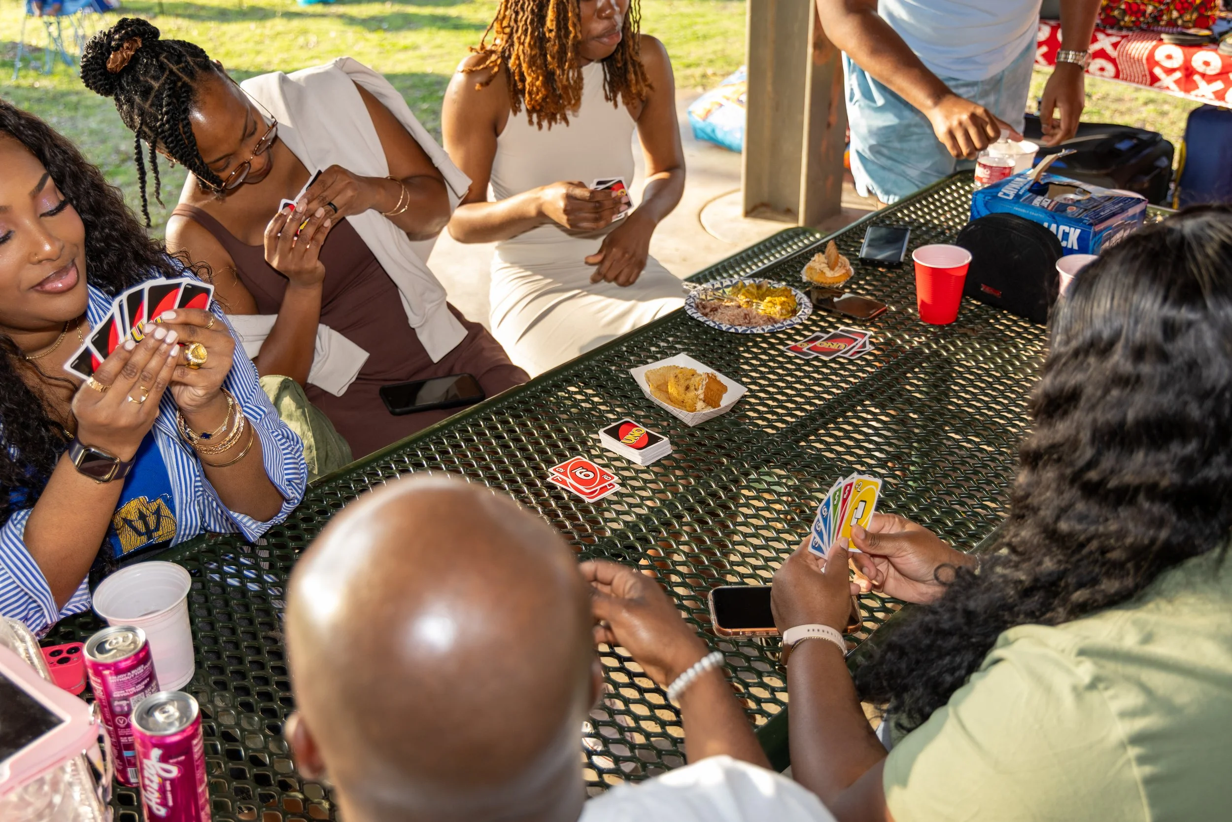 Group of people playing cards at an outdoor gathering, with snacks and drinks on a green metal table.