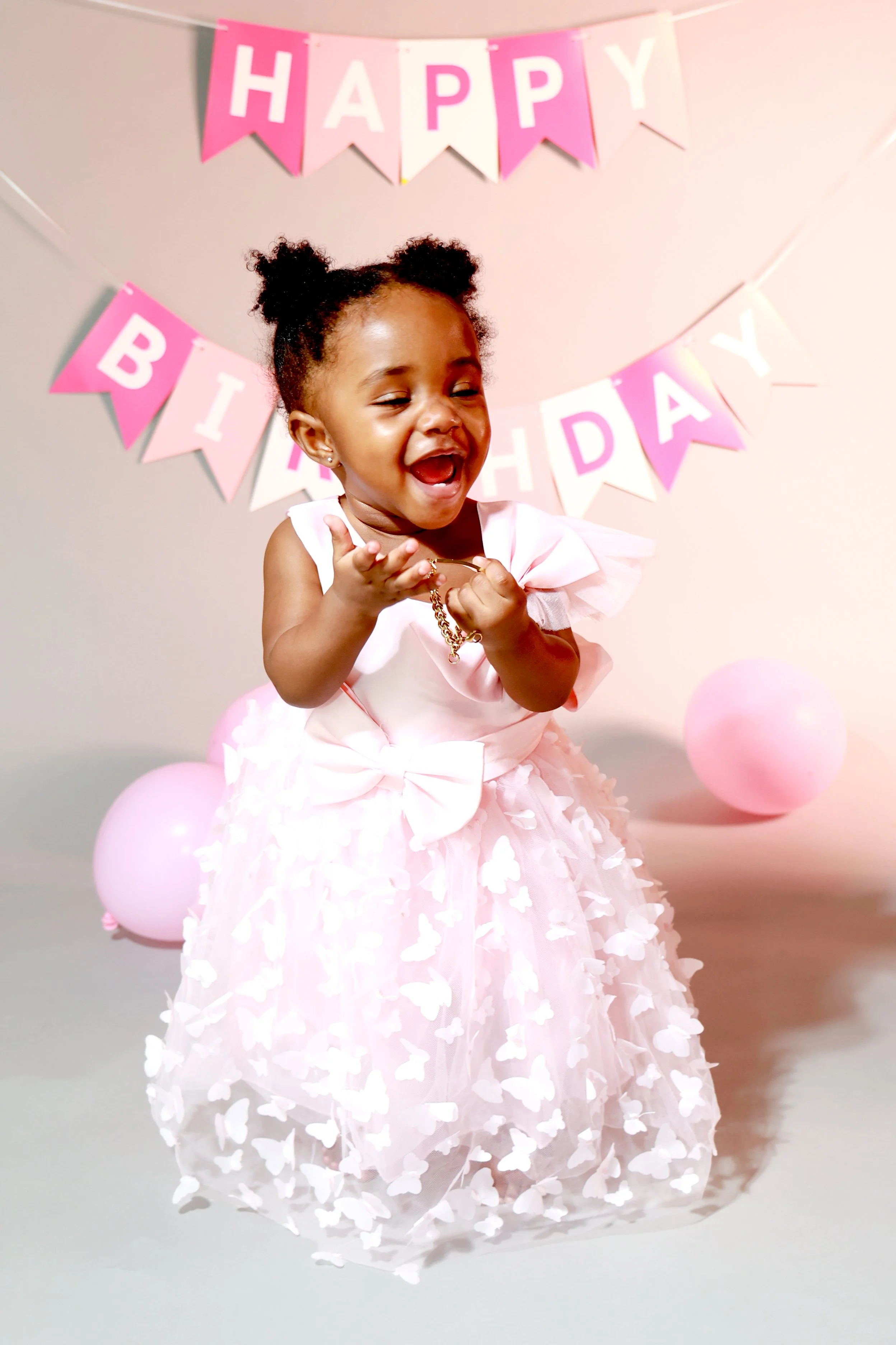 A young girl in a pink dress celebrating her birthday with pink balloons and a birthday banner that reads 'HAPPY BIRTHDAY' in the background.