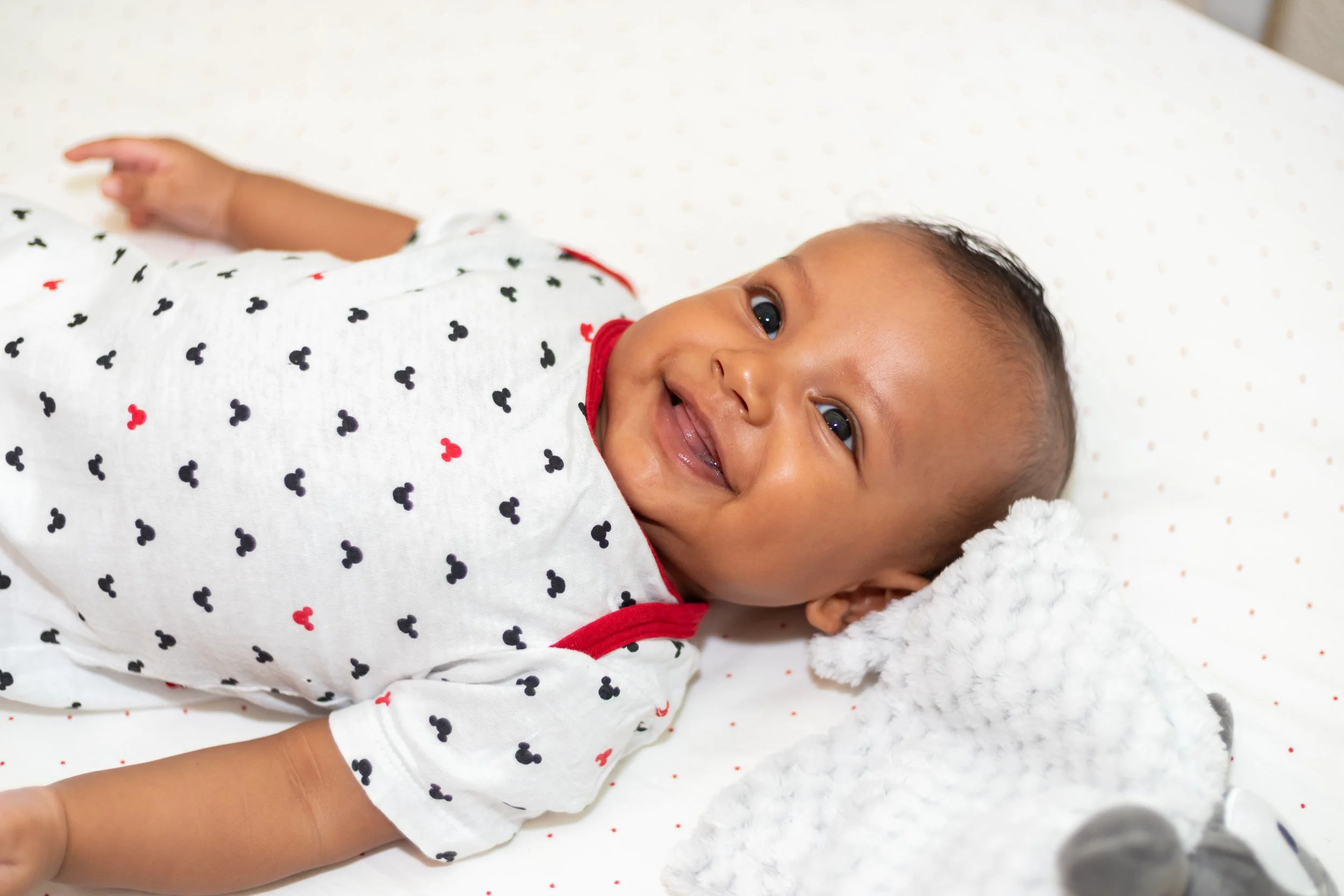 Smiling baby lying on a white polka dot blanket, wearing a white shirt with black and red Mickey Mouse print, and resting his head on a white plush toy.