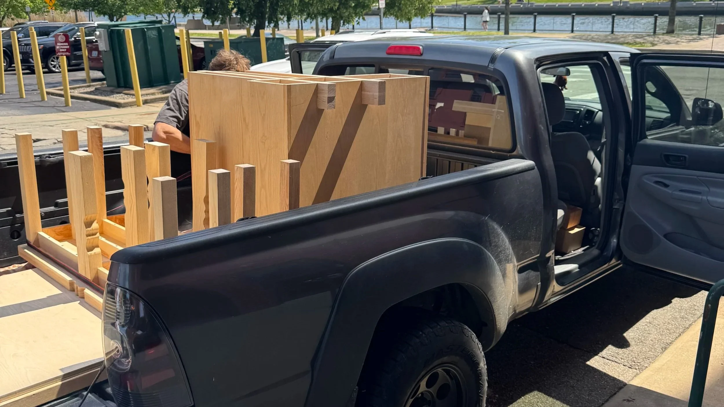 A black pickup truck parked in a parking lot with the passenger side door open, carrying wooden furniture or construction components in the truck bed.