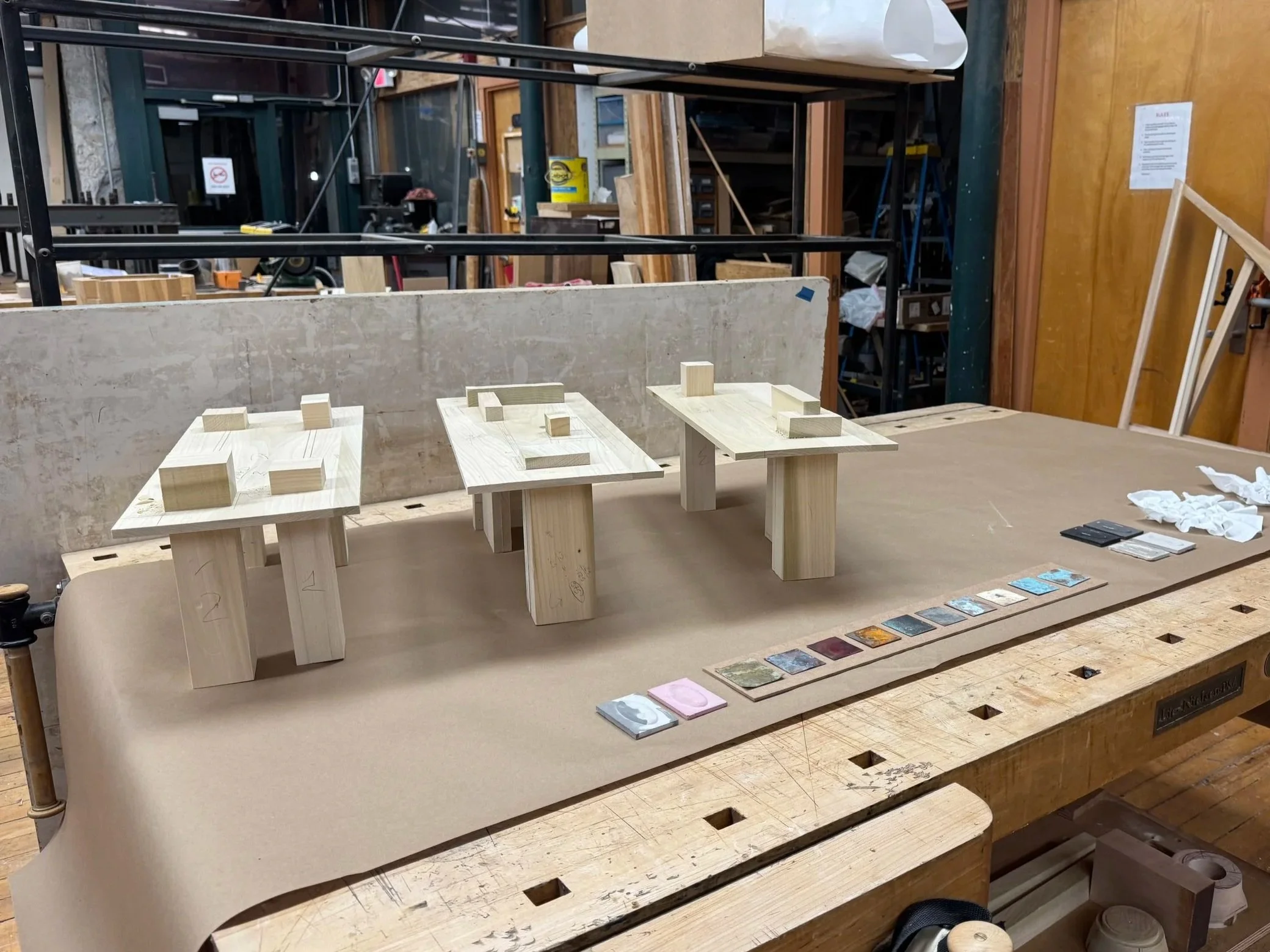 Wooden model of a building structure with three small wooden blocks and paint color samples on a workbench in a woodworking shop.