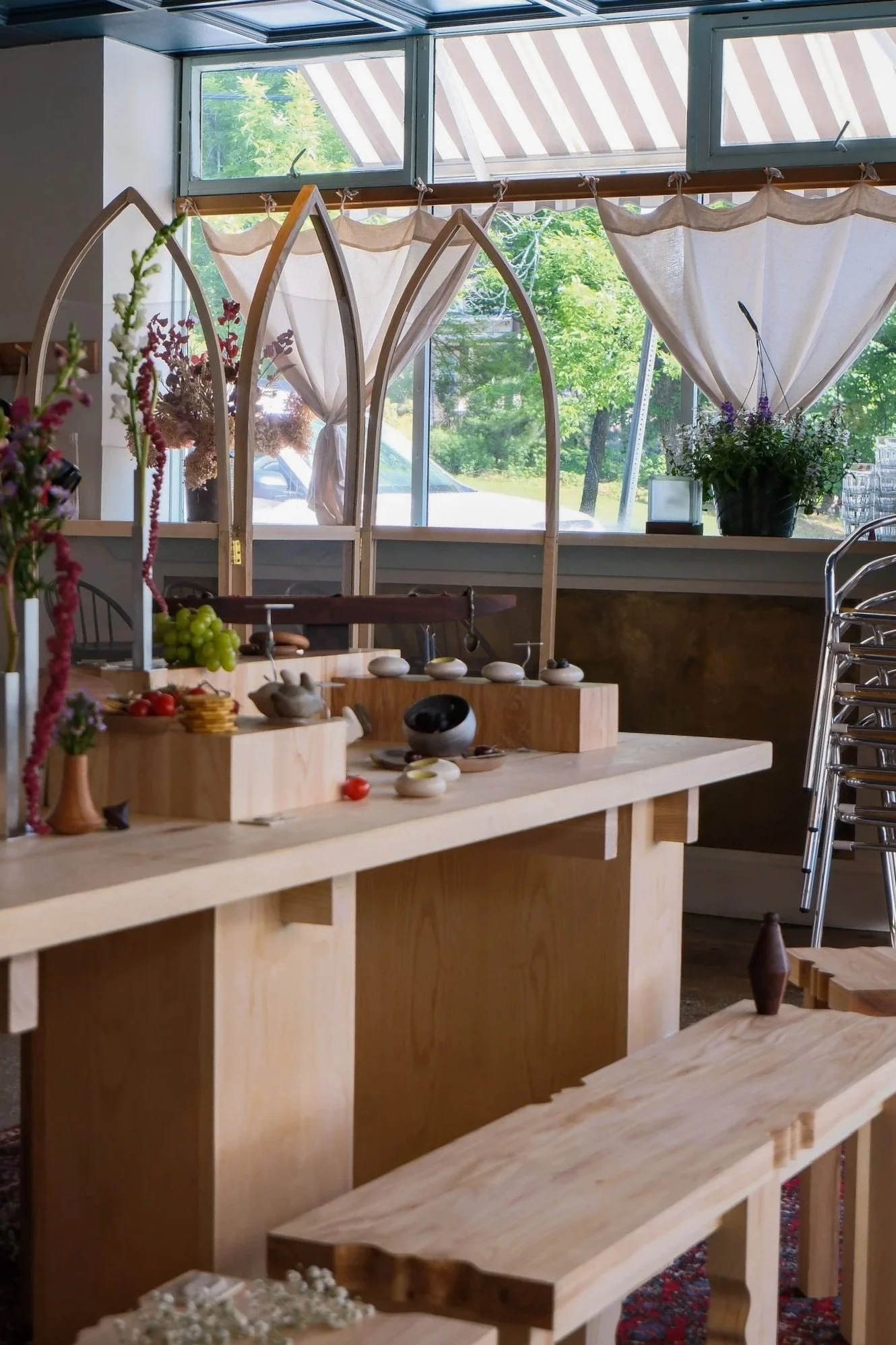 Inside a shop with wooden display tables and flower arrangements, large windows with curtains, and a view of trees outside.