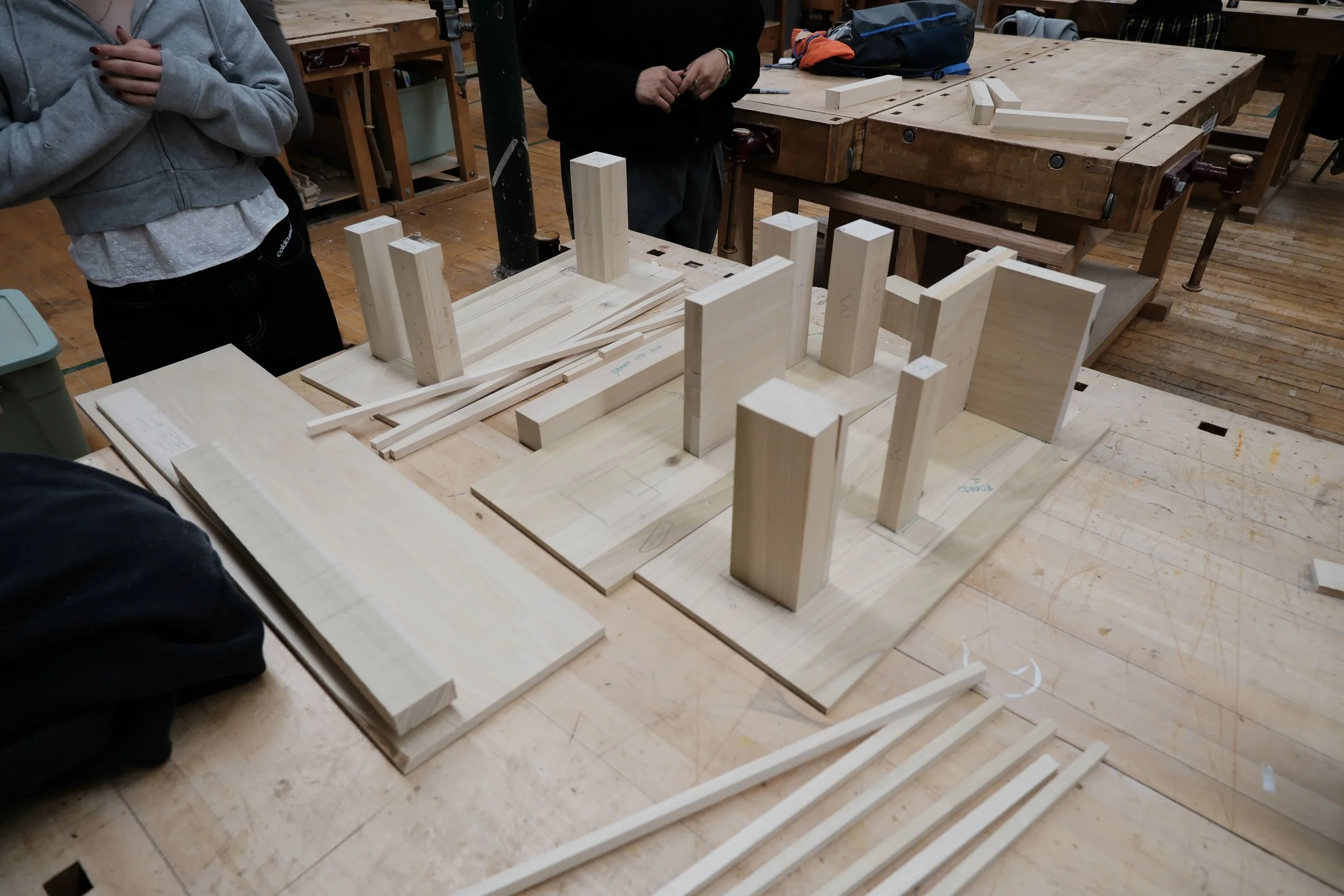 Wooden pieces and blocks arranged on a worktable in a woodworking shop, with people standing nearby.