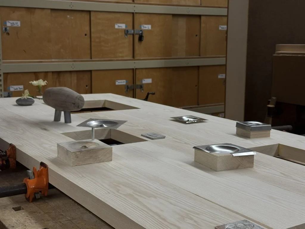 Workshop table with cutouts and metal dishes attached, set in a carpentry or woodworking shop with wooden cabinets in the background.
