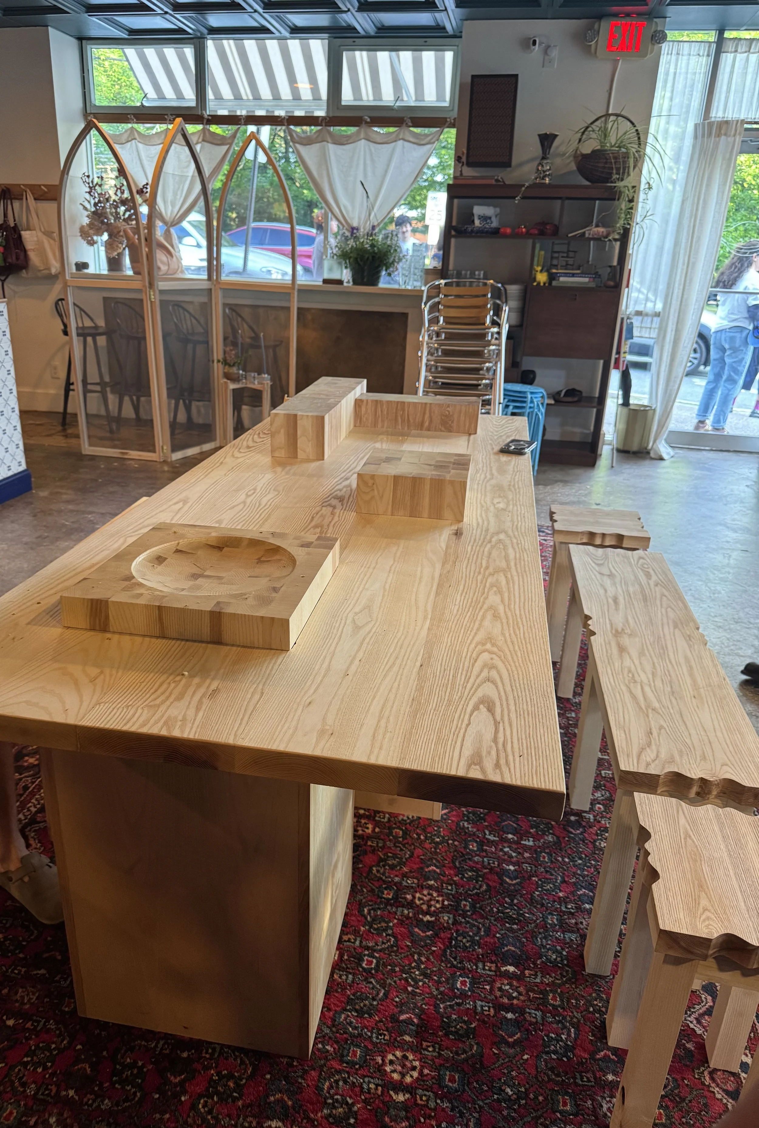 Interior of a cozy cafe with wooden furniture, including a large table with cutting boards, a shelving unit with decorative items, stacked chairs, and a window with curtains and plants outside.