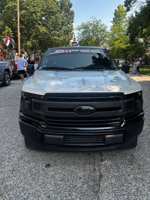 Front view of a black and silver Ford truck parked on a street during a gathering with people around.