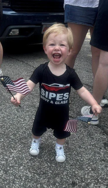 Small blonde boy smiling and holding two American flags, standing on a paved street with people in the background.
