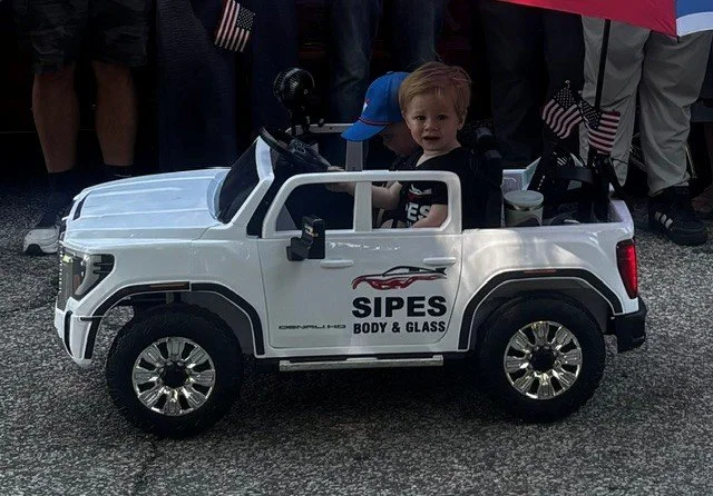 Child sitting in a miniature white electric toy truck with logo 'SIPES Body & Glass' on the side, surrounded by people and holding an umbrella.