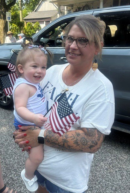 A woman holding a young girl, both smiling and holding small American flags, standing outdoors near parked cars, with trees and houses in the background.