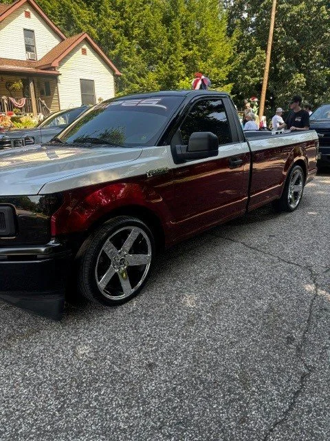 A pickup truck with a two-tone paint job, chrome wheels, and a black hood at a local event, with people gathered in the background and a house nearby.