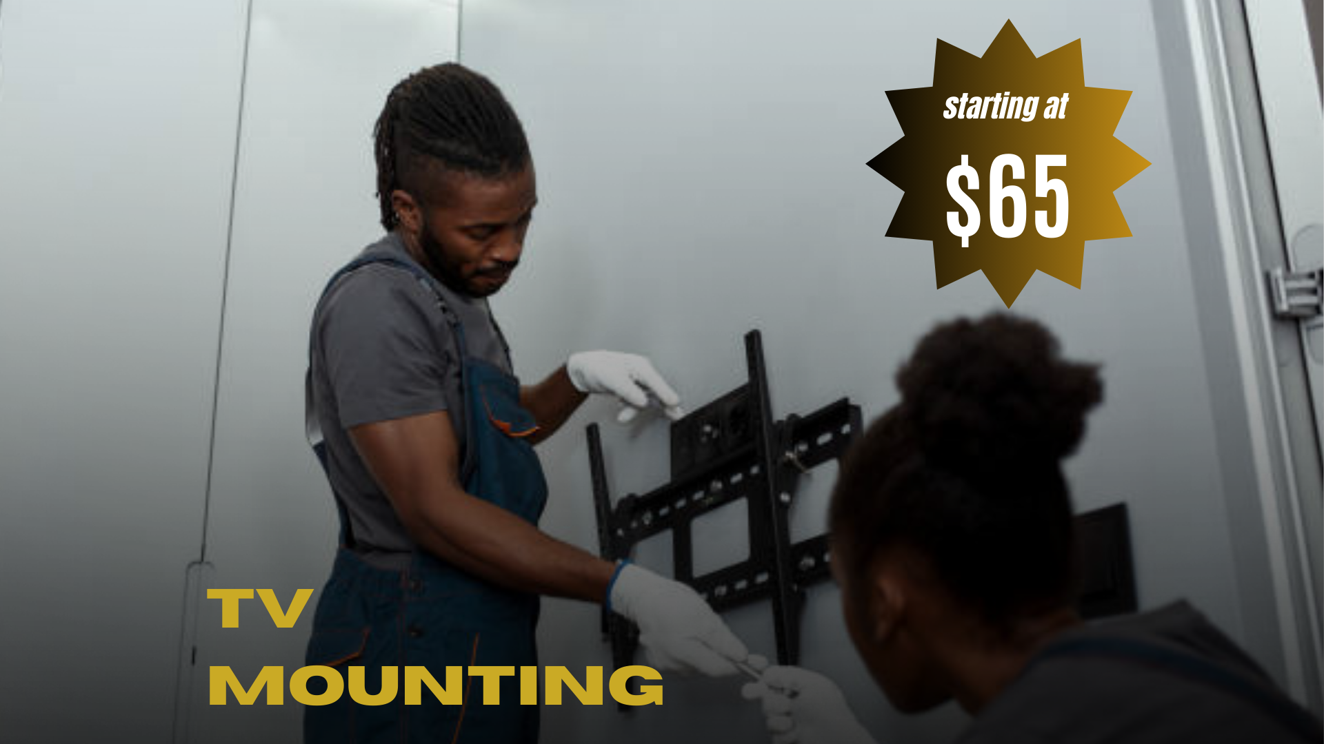 Two people mounting a TV, one standing and one kneeling, with a wall mount and a TV bracket in a room with plain gray walls.