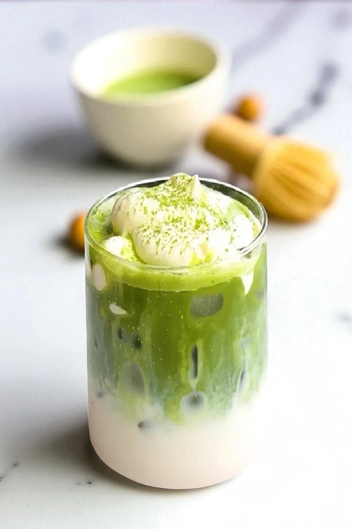 A glass of matcha latte topped with whipped cream, with a bowl of matcha in the background.