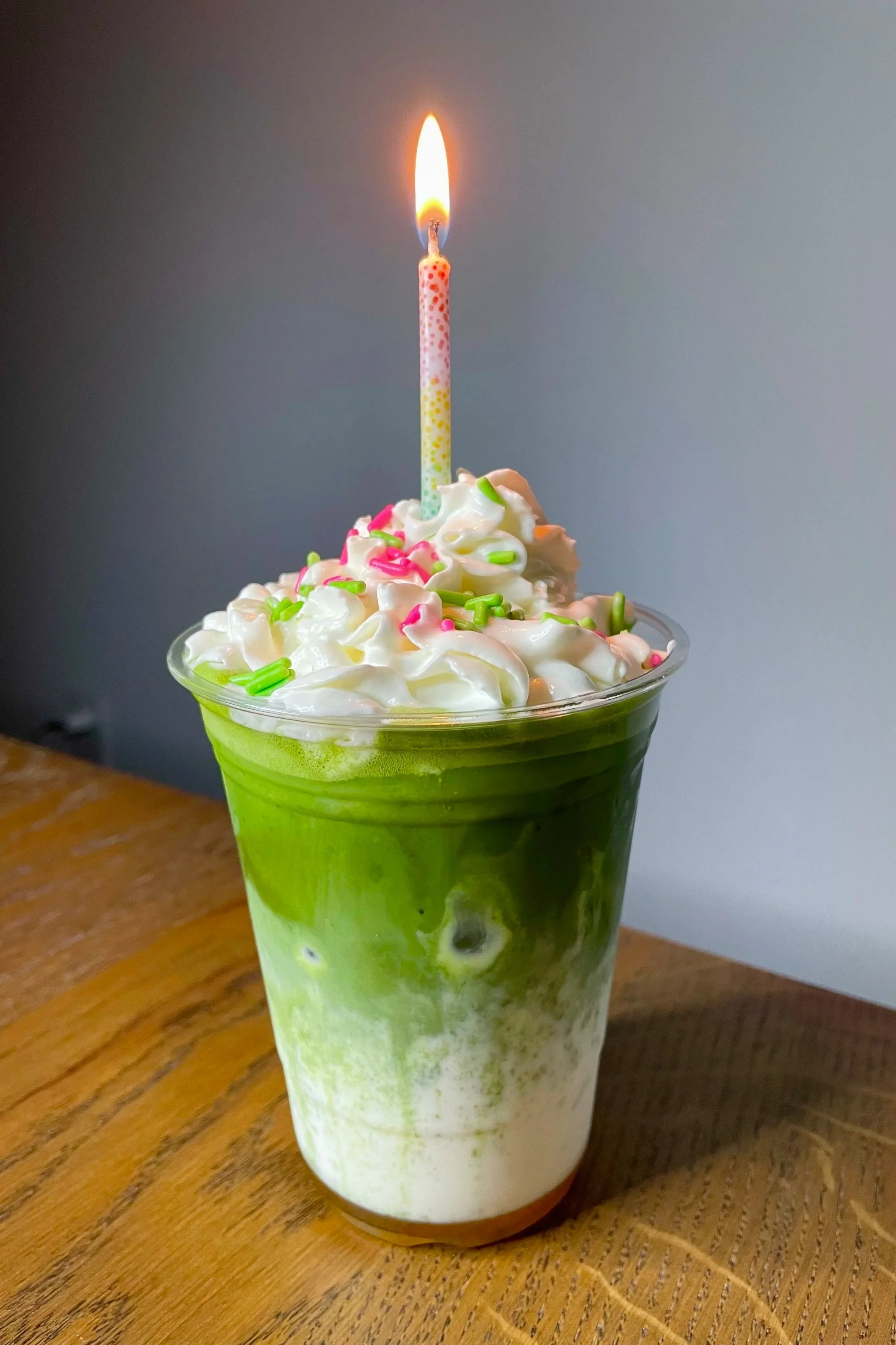A green and white layered iced coffee or matcha drink in a clear cup topped with whipped cream, colorful sprinkles, and a lit birthday candle.