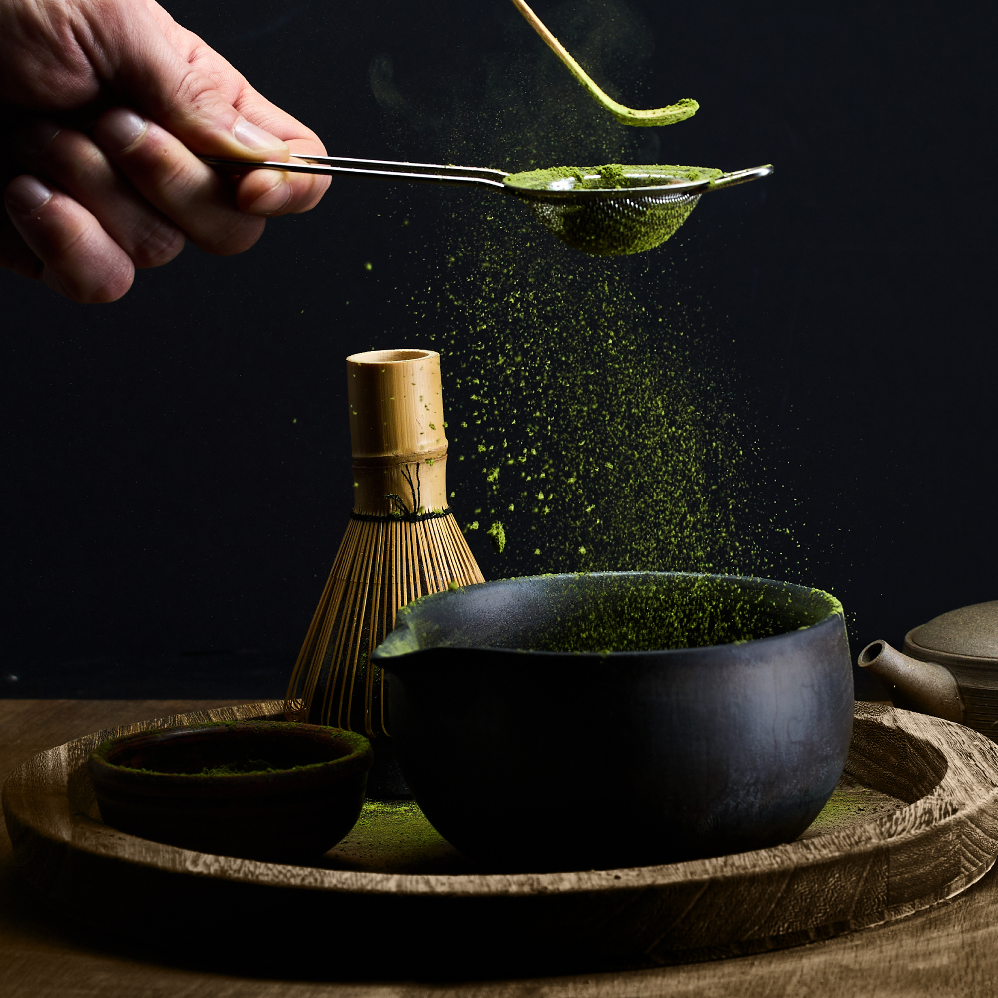 A hand sifting matcha green tea powder through a fine sieve into a black bowl, with traditional tea utensils on a wooden tray.