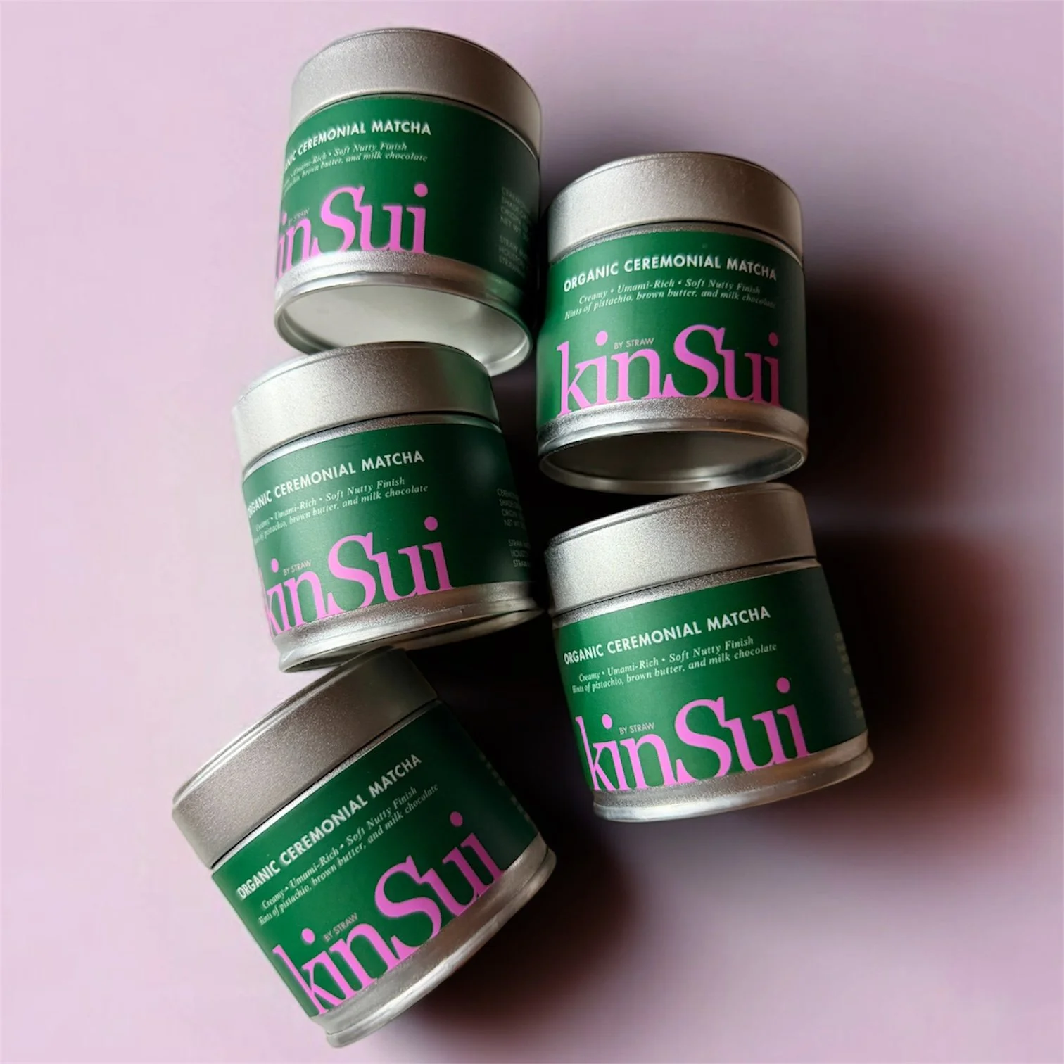 Six small cans of kinsui organic ceremonial matcha tea with green labels and pink text, arranged on a pink background.