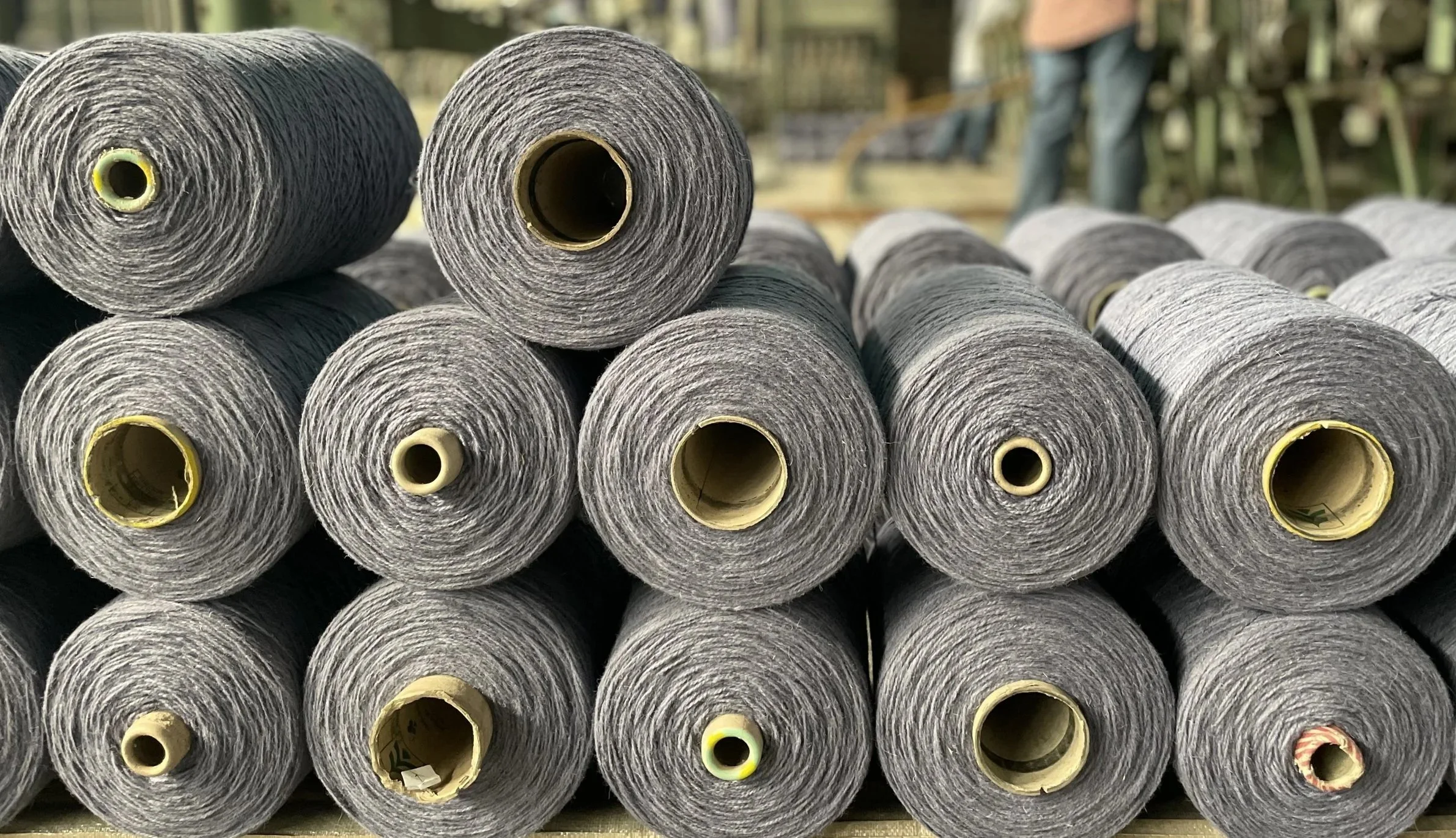 Multiple gray yarn spools stacked in a pyramid at a store or warehouse with a blurry person in the background.