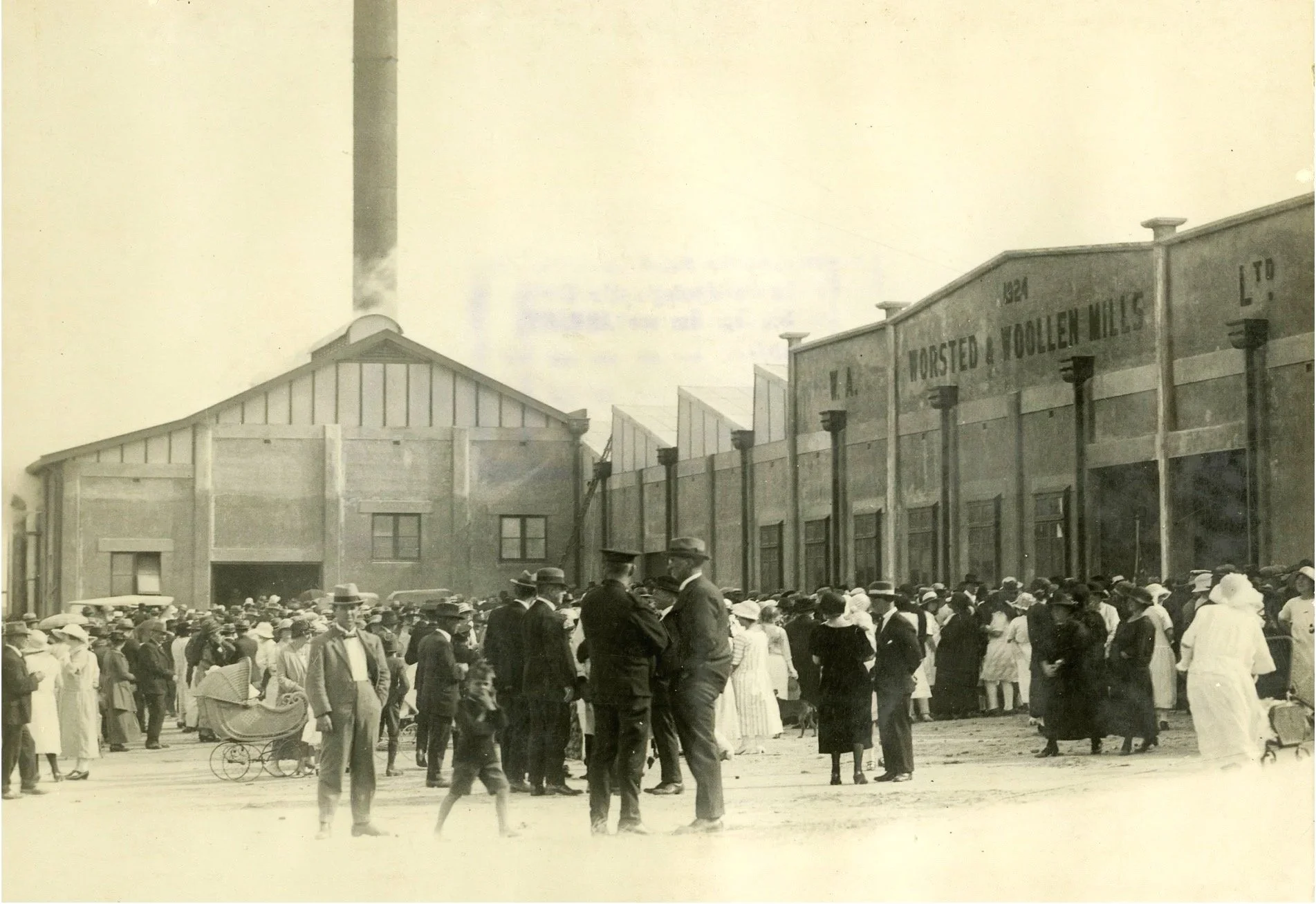 Historical black and white photo of a crowd of people in front of industrial buildings with a chimney in the background. People are dressed in early 20th-century clothing, including men in suits and hats, women in dresses and hats, and children. Some are standing, walking, or pushing a baby carriage.