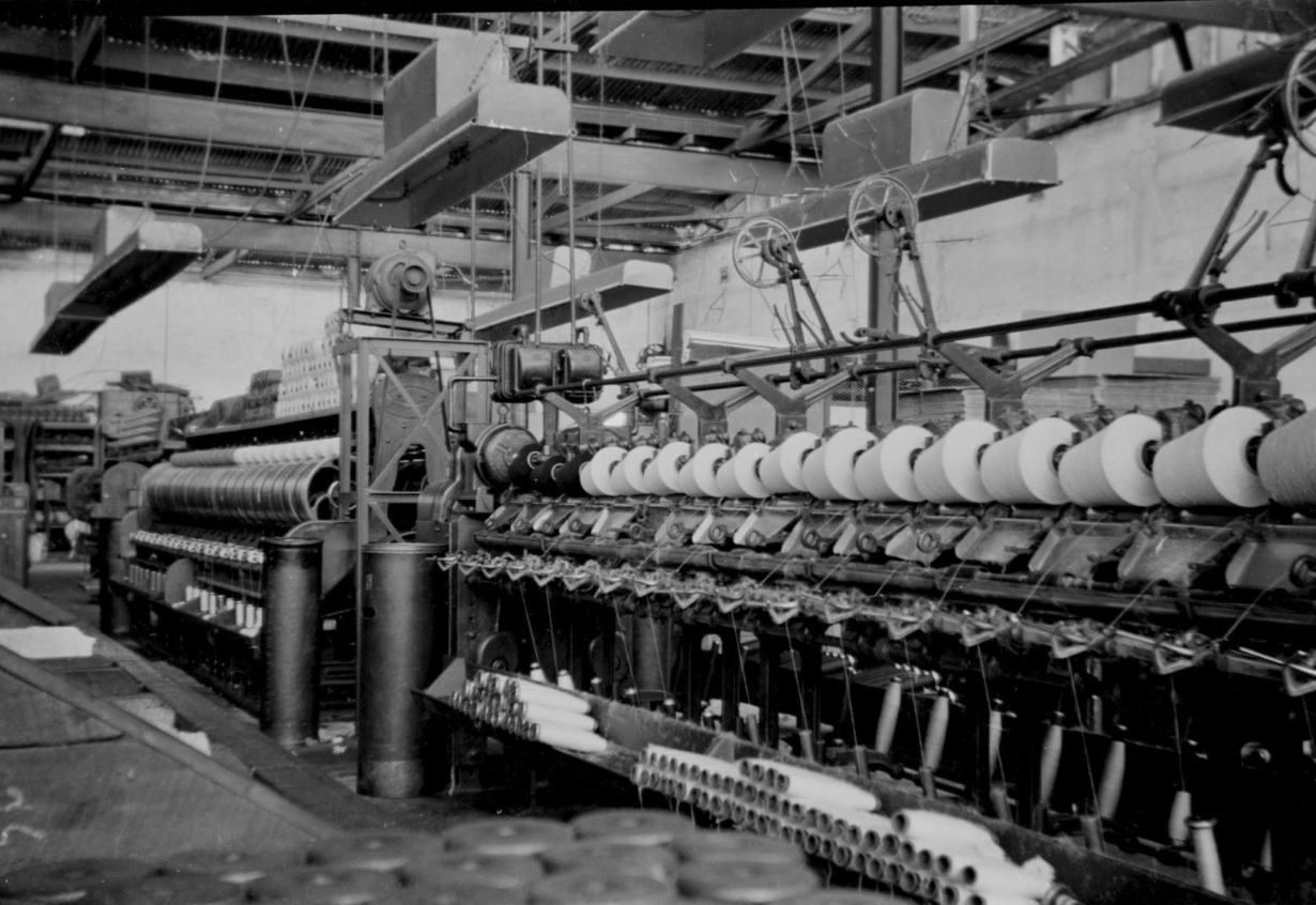 Black and white photo of an industrial manufacturing machine in a factory setting, with rollers, spools, and mechanical parts involved in textile or fabric production.