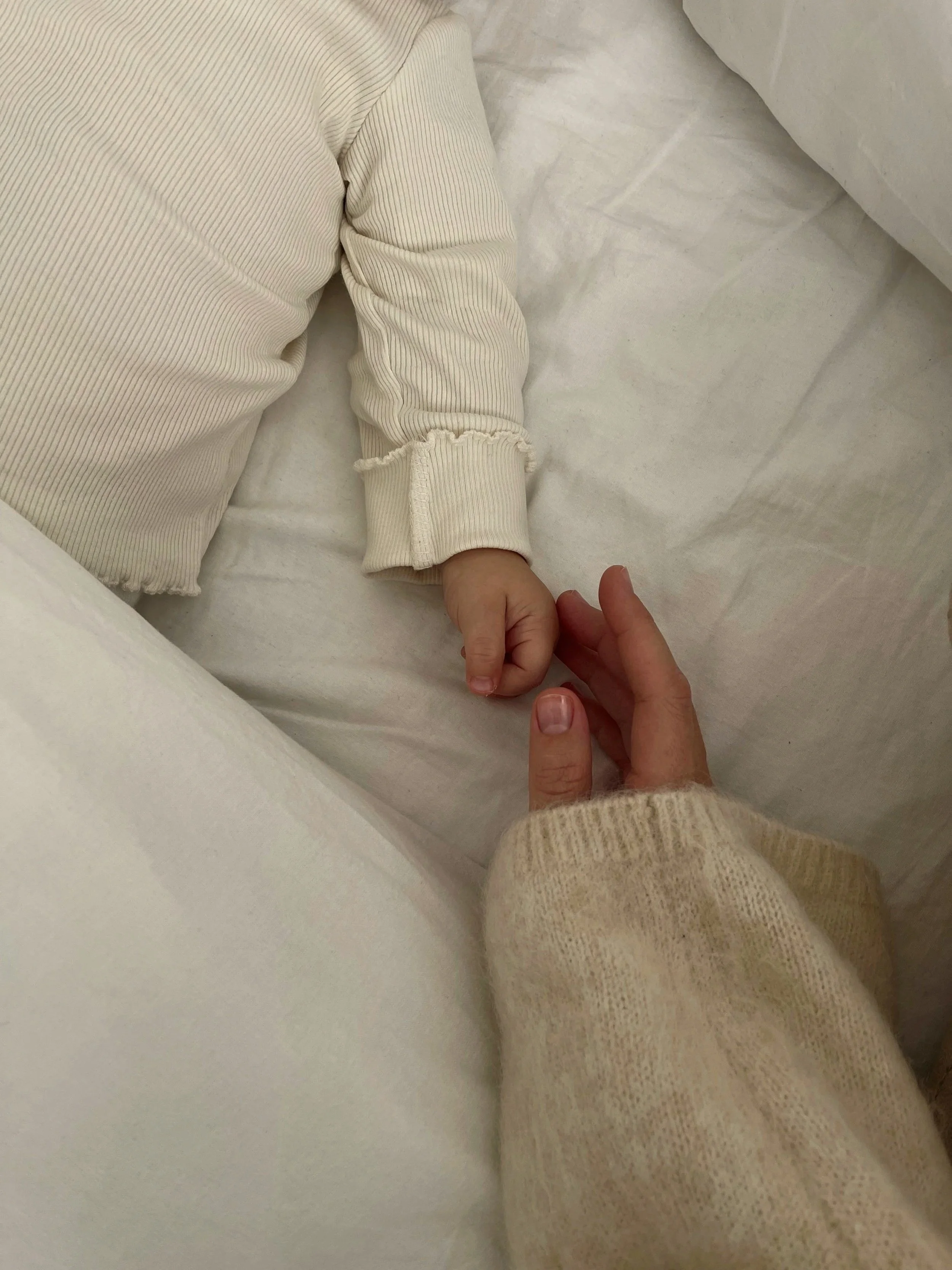 A close-up of an adult hand gently holding the tiny finger of a baby lying on a white bed. The baby is wearing a long-sleeved, beige, ribbed onesie with ruffled cuffs.