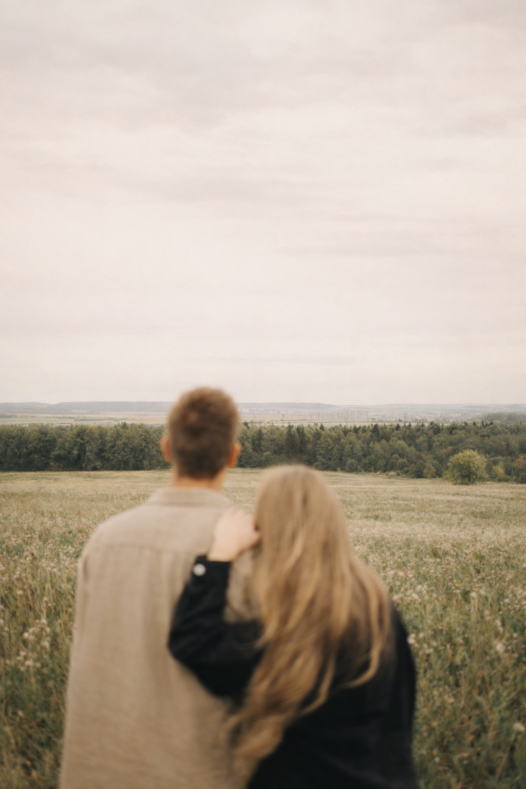 A couple stands in a field overlooking a distant horizon with trees and open land, viewed from behind.