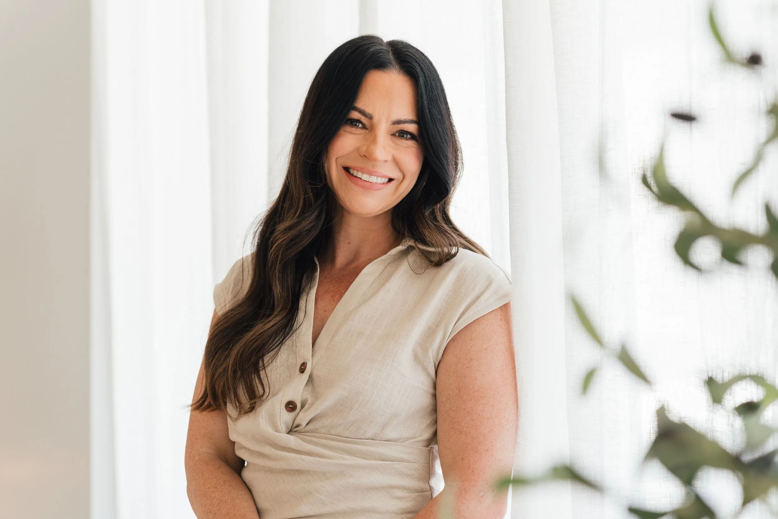 A woman with long, dark, wavy hair smiling and looking slightly to the left in front of a light curtain with some green leaves in the foreground.