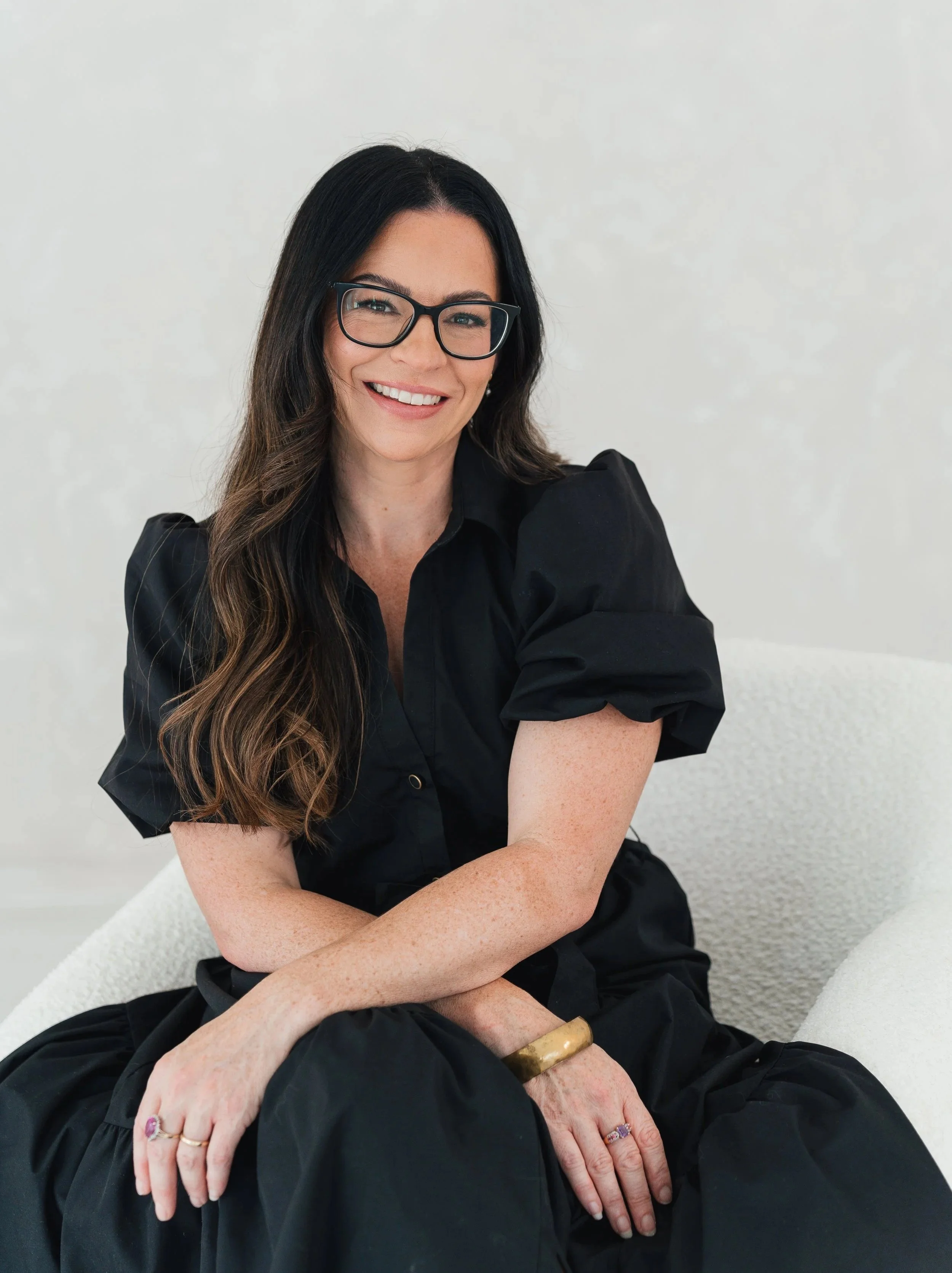 A woman with long, dark hair, wearing glasses and a black shirt, smiling and sitting on a white textured chair, with her arms crossed and wearing multiple rings and a gold bracelet.