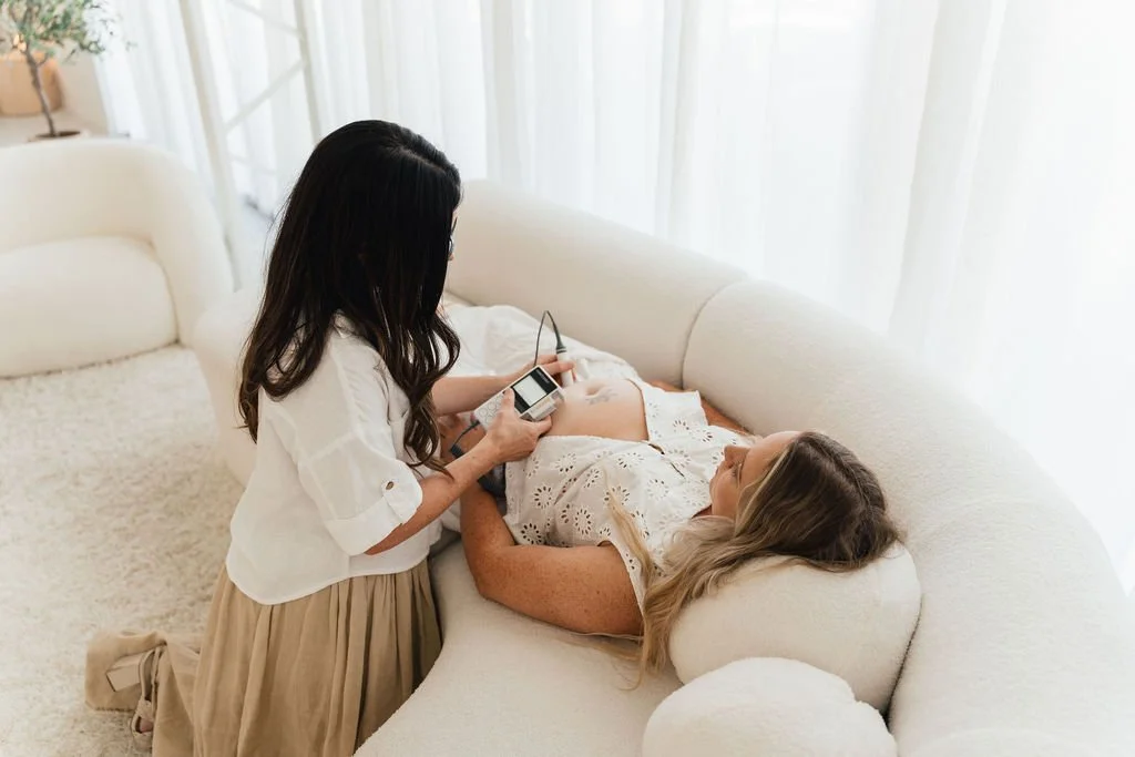 A woman lying on a white sofa, with a blanket, using a baby heartbeat monitor, while another woman stands beside her, holding a device connected to the monitor, in a bright living room.