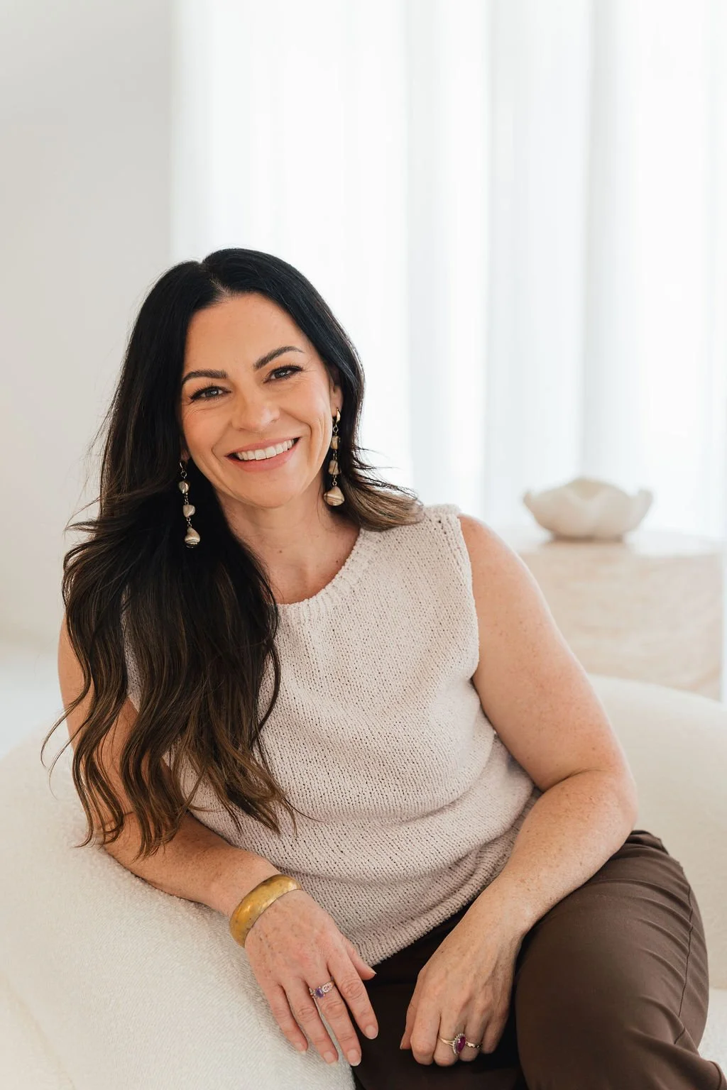 A woman with long dark hair, smiling, wearing a sleeveless beige top, and earrings, sitting on a cream-colored couch in a bright room with white curtains and decorative bowl in the background.