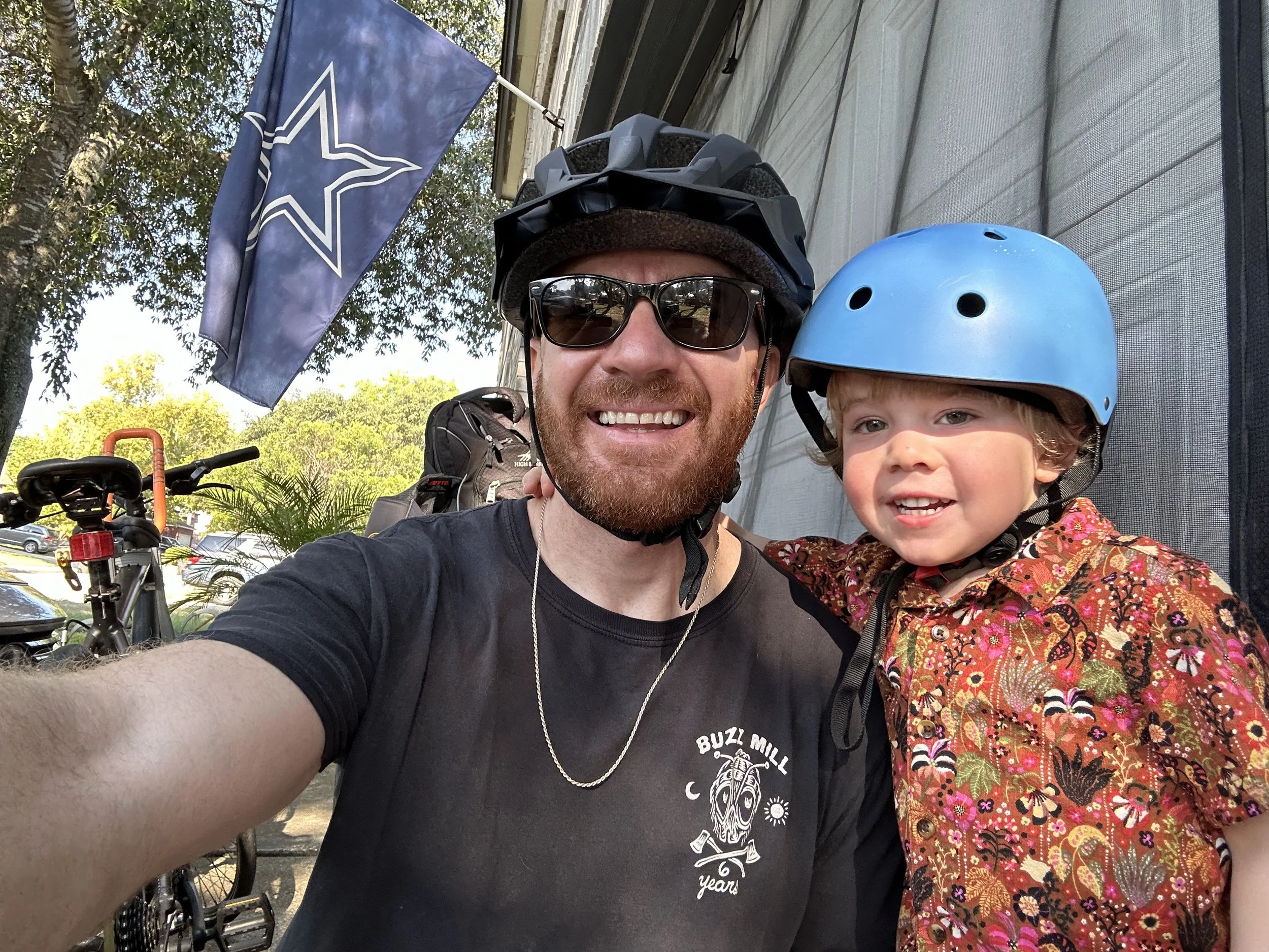 A man and a young both, both wearing bike helmets, taking a selfie outdoors on a sunny day. There are trees, bikes, parked cars, and a Dallas Cowboys flag in the background.