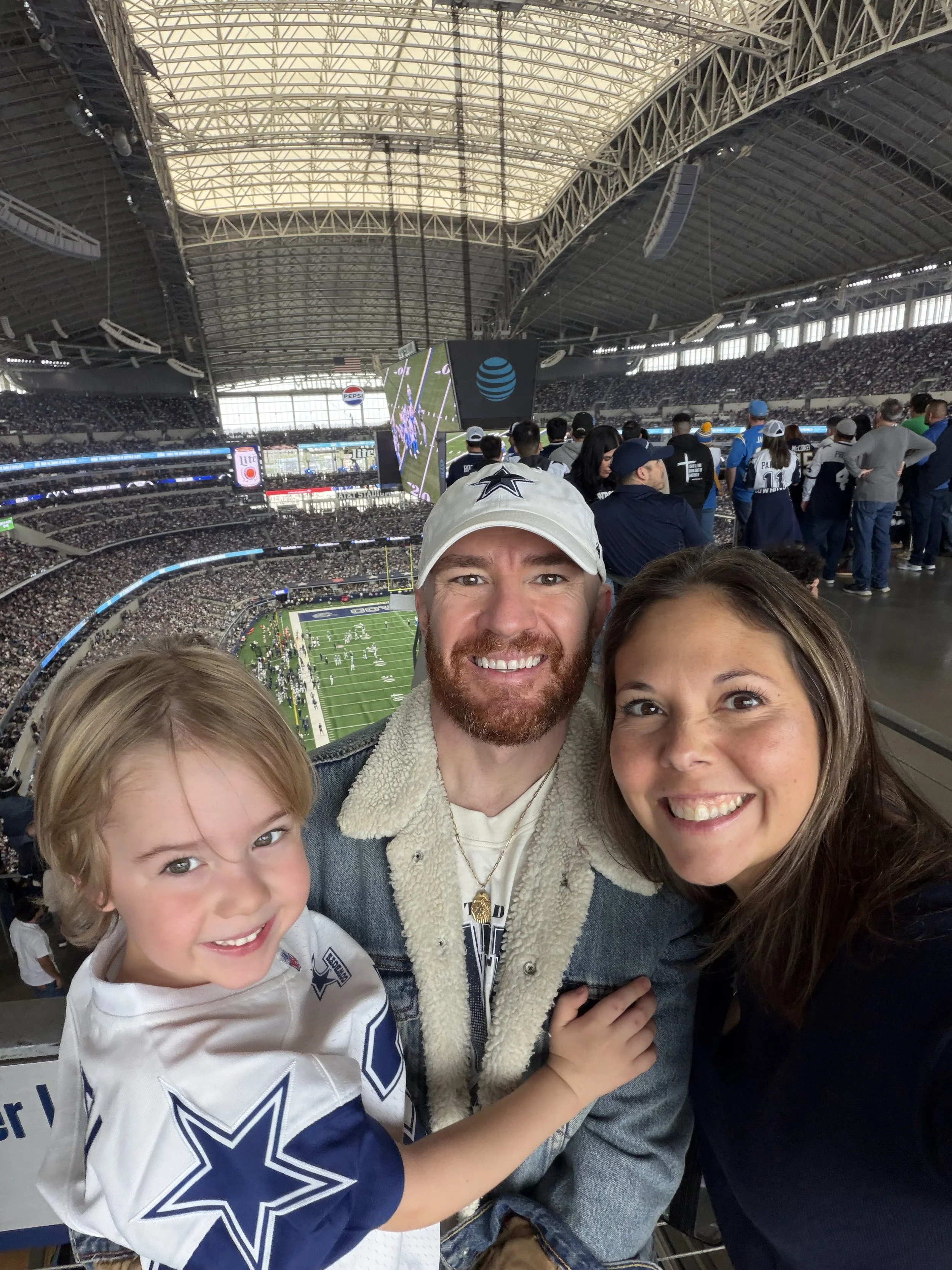 Three people, including a young boy and two adults, smiling and posing for a selfie inside a large, enclosed stadium during a football game. The stadium is filled with spectators, and a big screen displays the game.