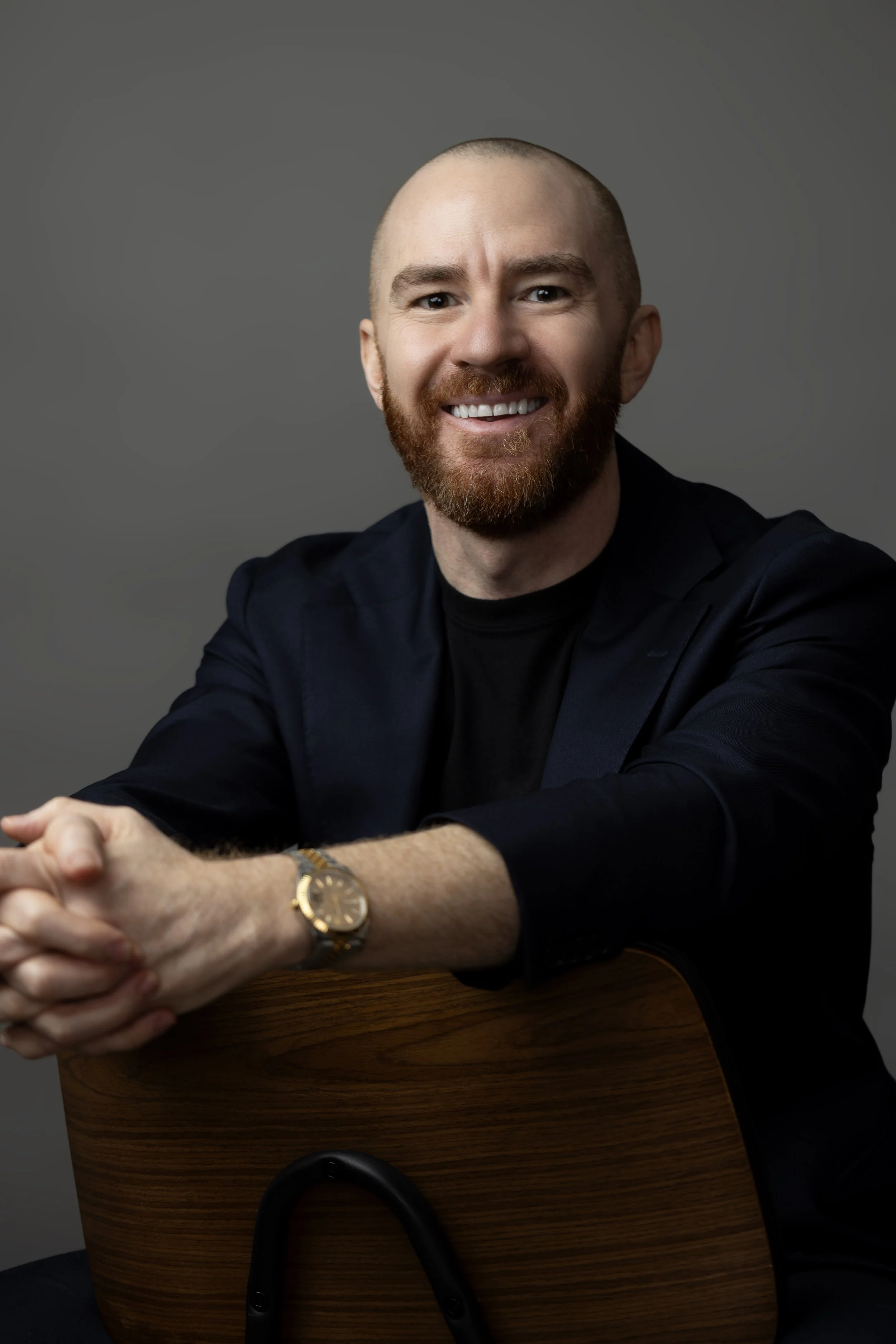 A man with a shaved head and red beard is smiling, sitting at a wooden table, wearing a black jacket and a gold wristwatch.