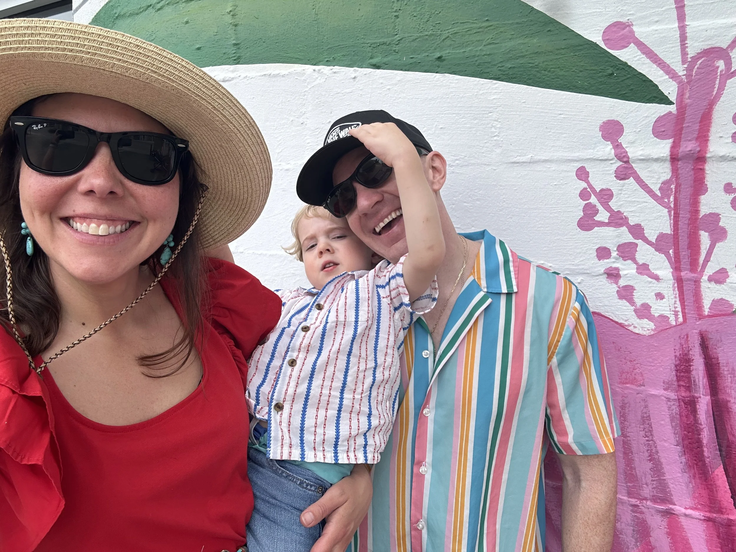A woman wearing a large straw hat and dark sunglasses smiling. A man in a black cap and sunglasses holding a young boy in colorful striped shirt, with a mural painted on a white wall in the background.